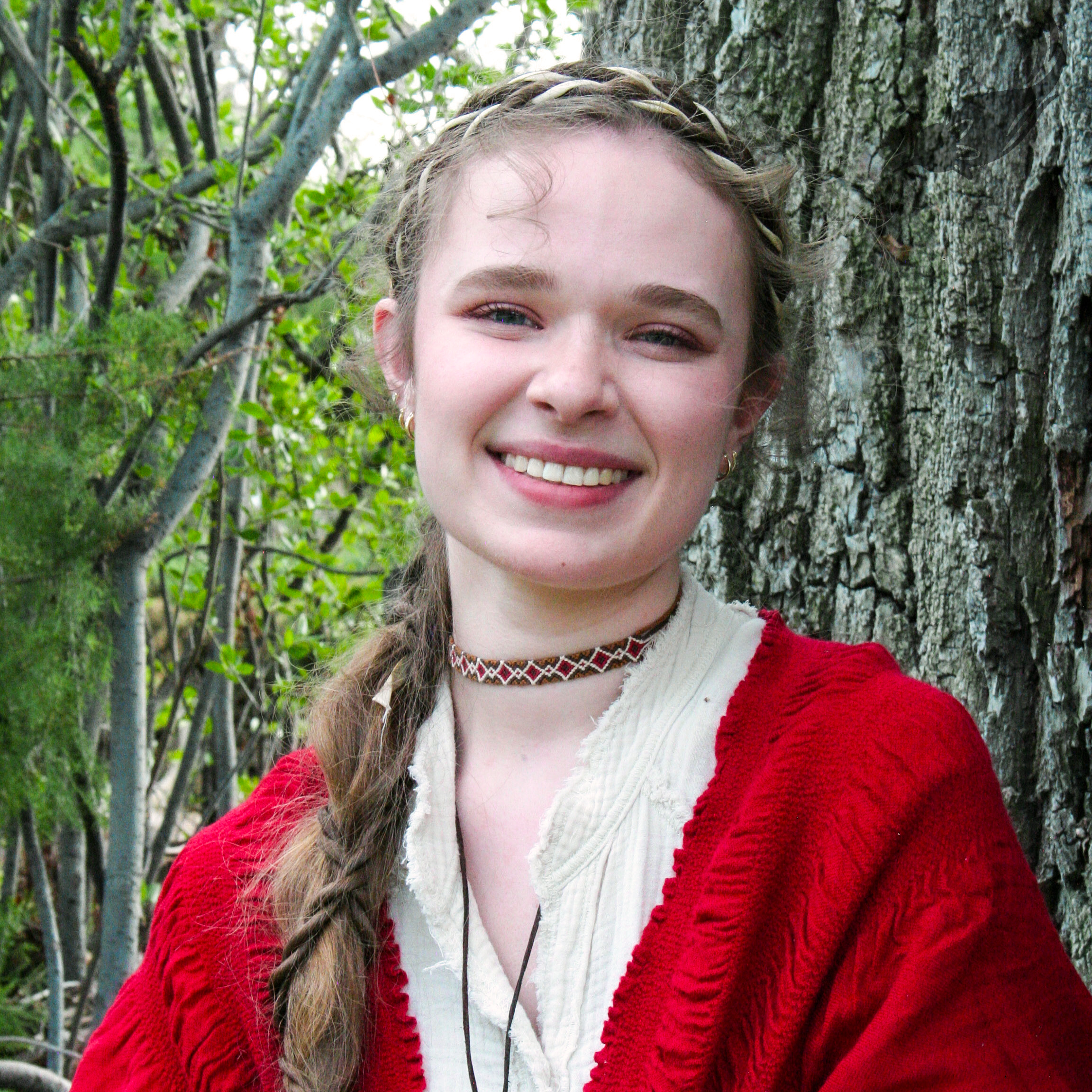 A smiling young woman with long hair and a braided headband standing in front of a tree with green leaves.