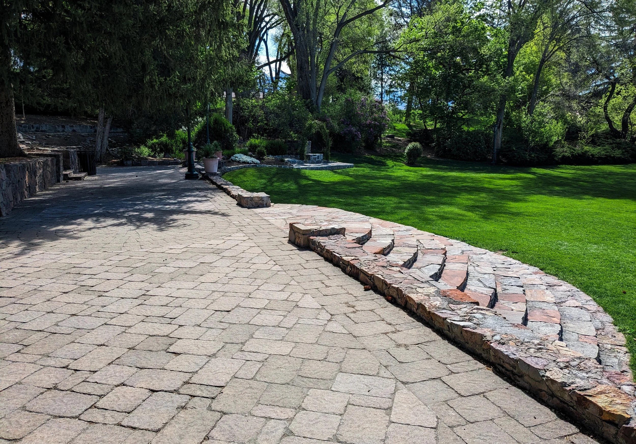 A paved garden pathway with a stone border on the left side, leading through a lush green backyard filled with trees and shrubs, with sunlight filtering through the foliage.