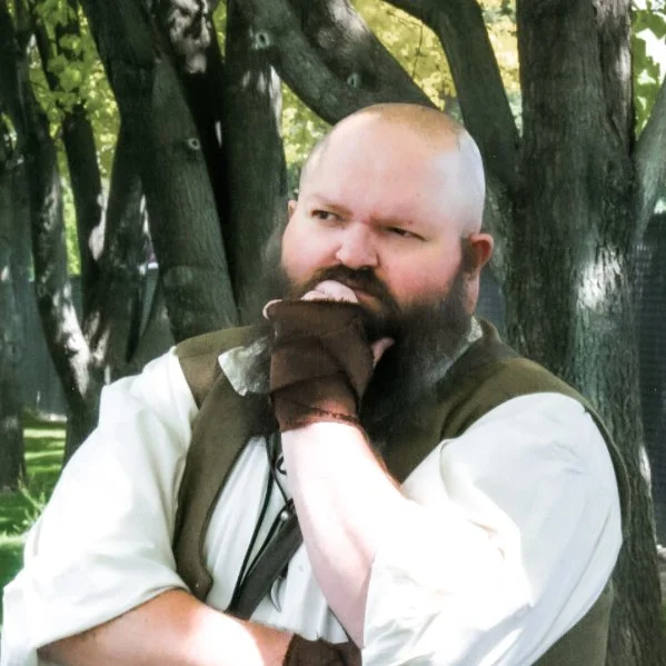 A man with a beard and a shaved head, wearing medieval or fantasy-inspired clothing, standing outdoors near trees, with a thoughtful expression and hand on his chin.