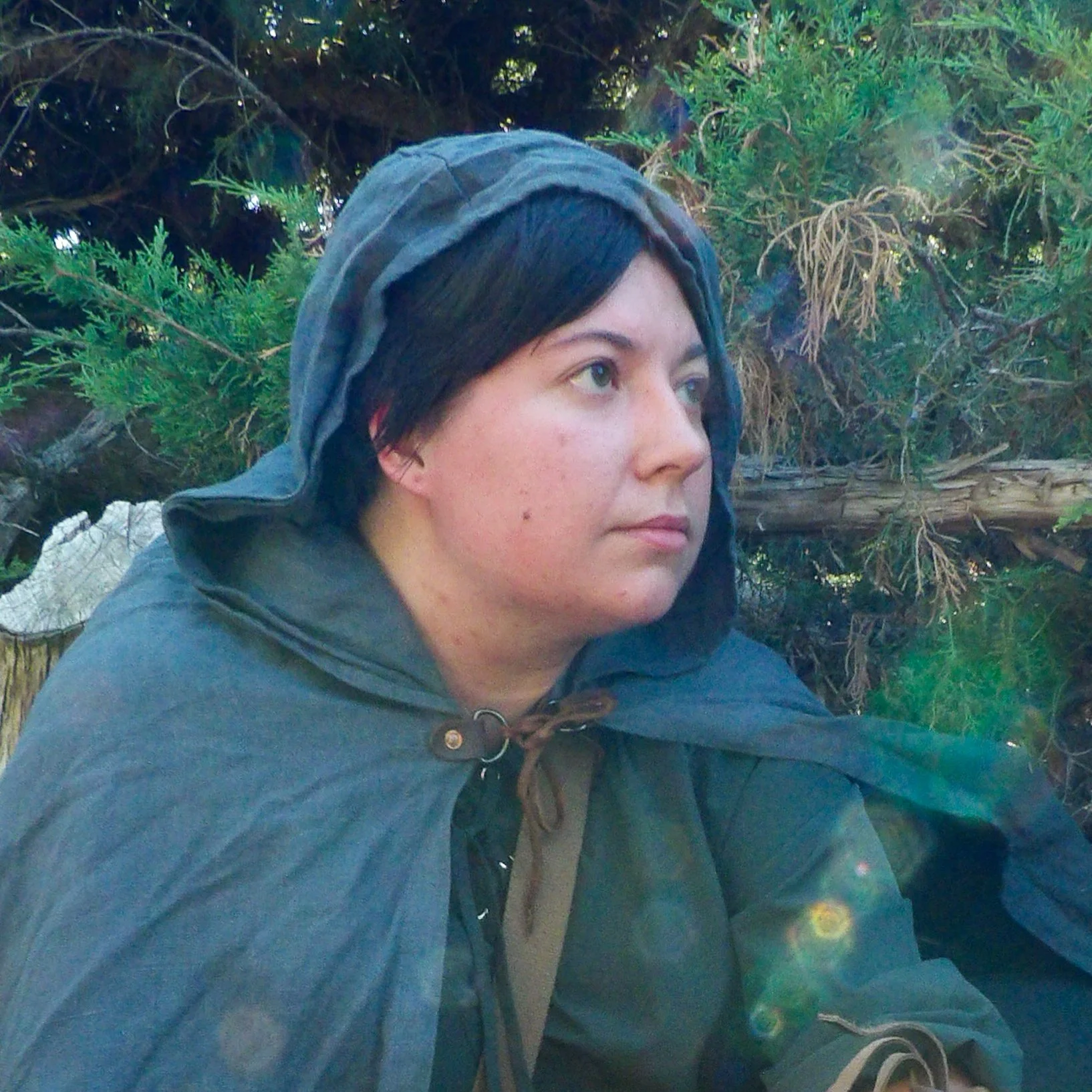 A person with short dark hair, wearing a blue hoodie, sitting outdoors among green bushes and trees, with sunlight creating lens flare effects.