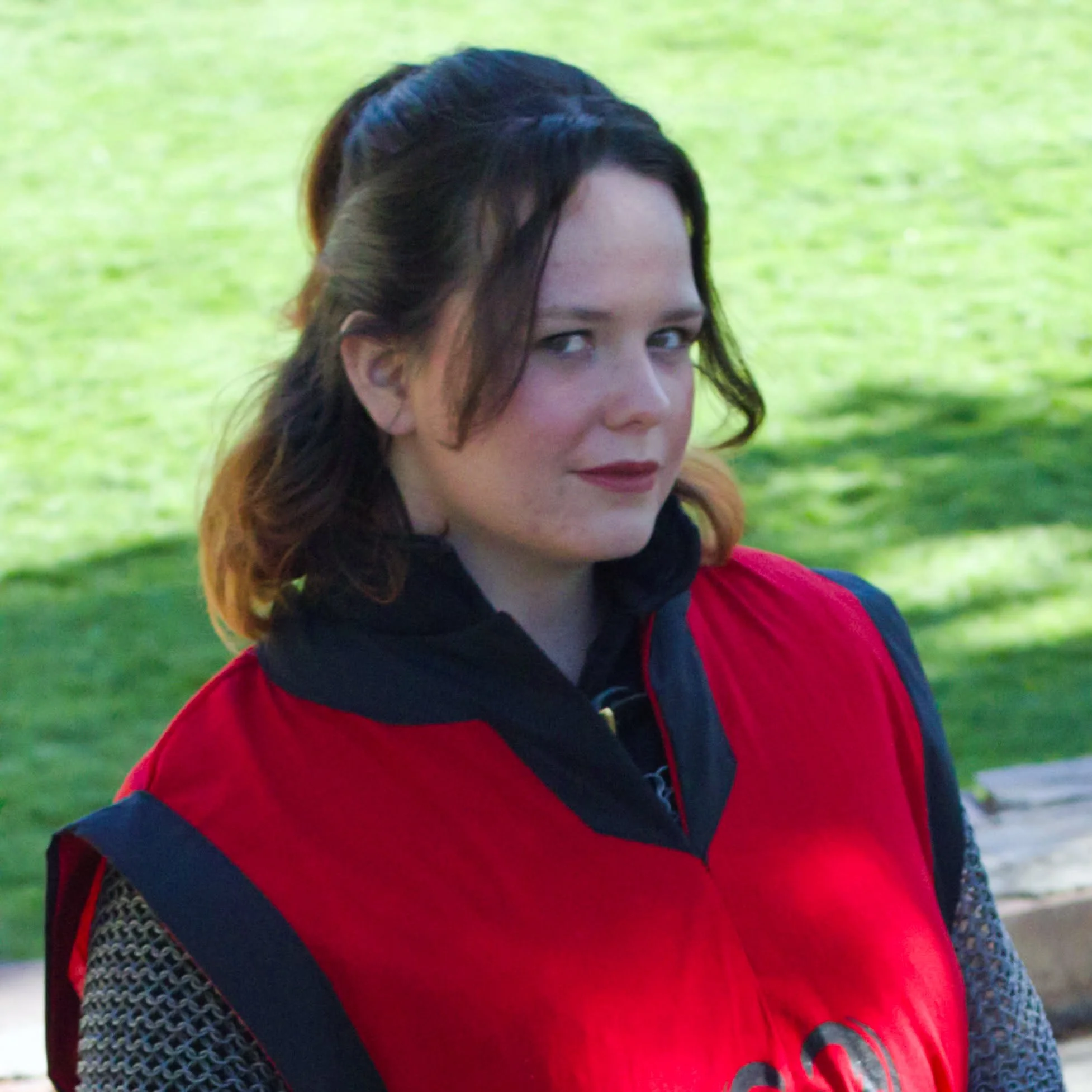 Close-up of a young woman with dark hair, wearing a red and black jacket, outdoors on a green grassy area.
