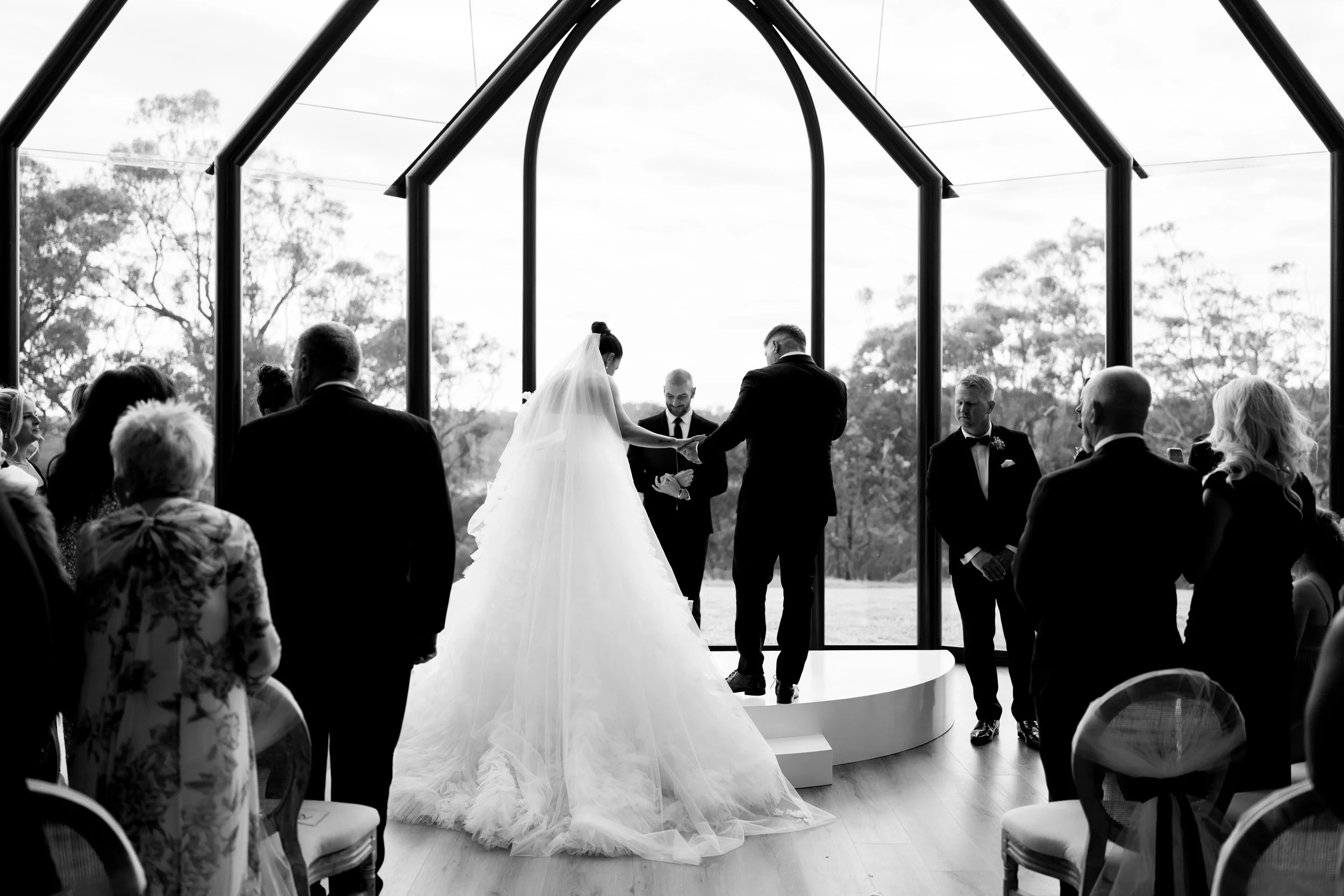 A black and white wedding ceremony inside a glass greenhouse with the bride and groom holding hands, surrounded by guests.