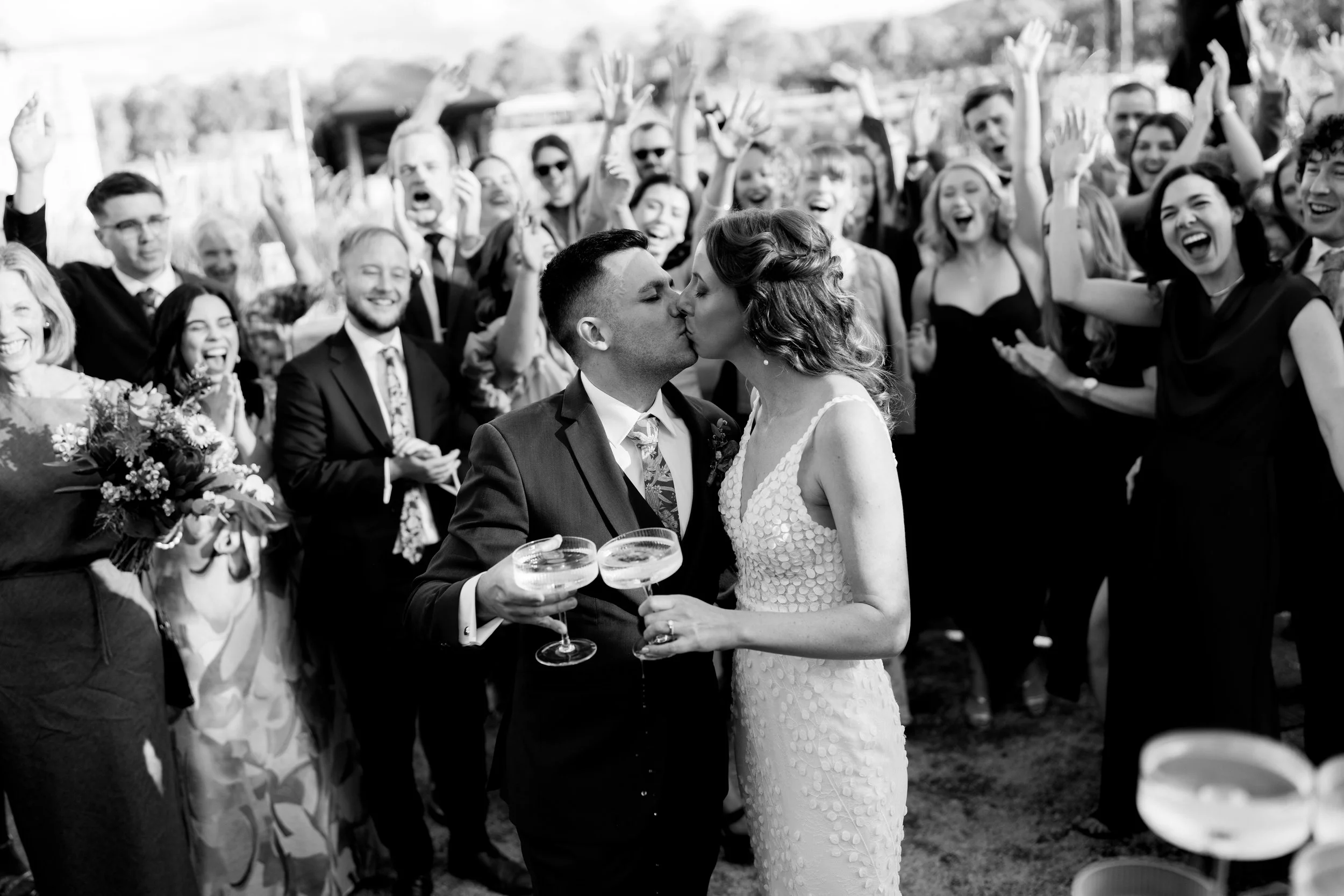 A newly married couple kissing at their wedding celebration surrounded by friends and family.