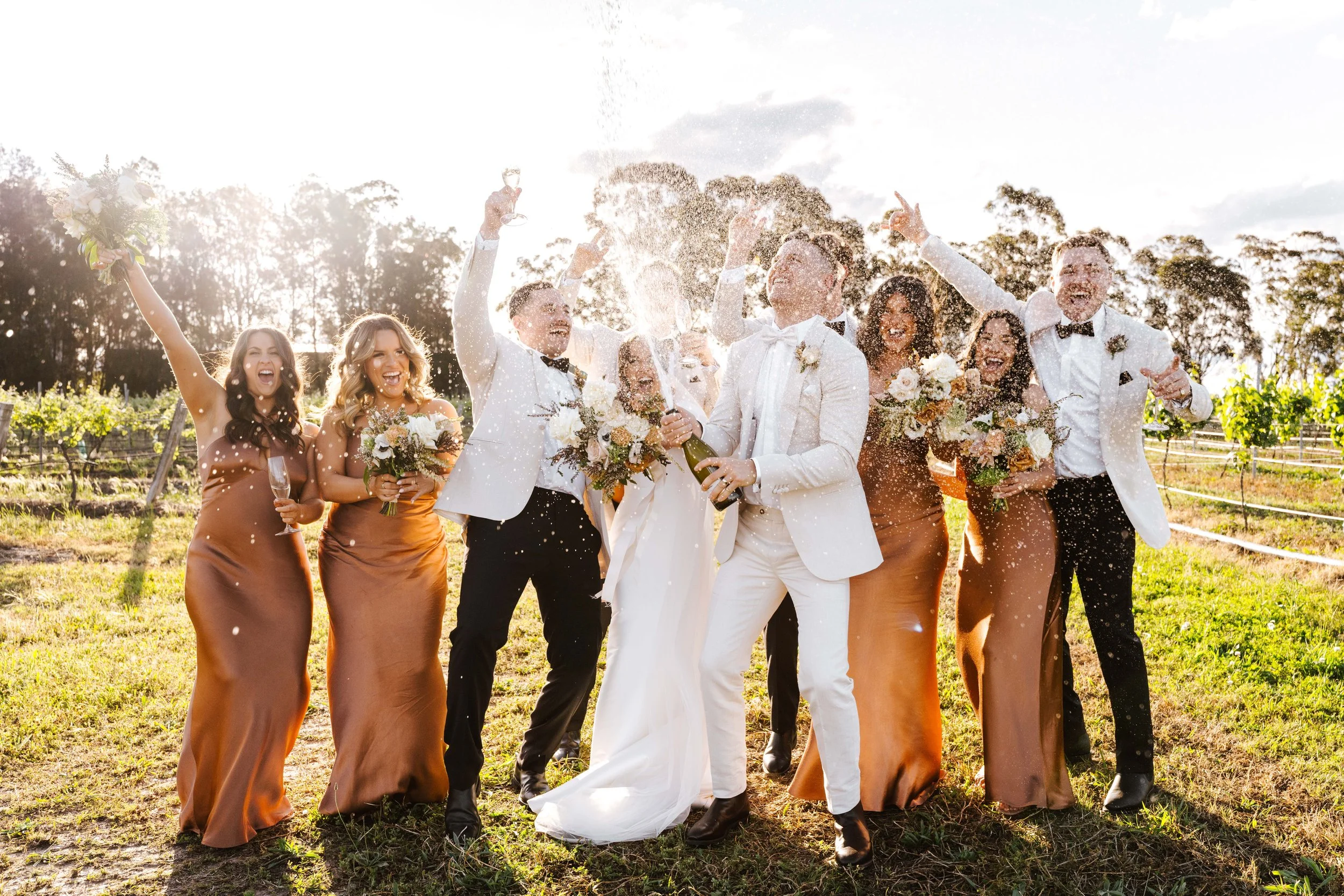 Wedding celebration with the bride, groom, and wedding party outdoors in a vineyard, decked out in formal attire, celebrating with champagne and floral bouquets as champagne spray is thrown