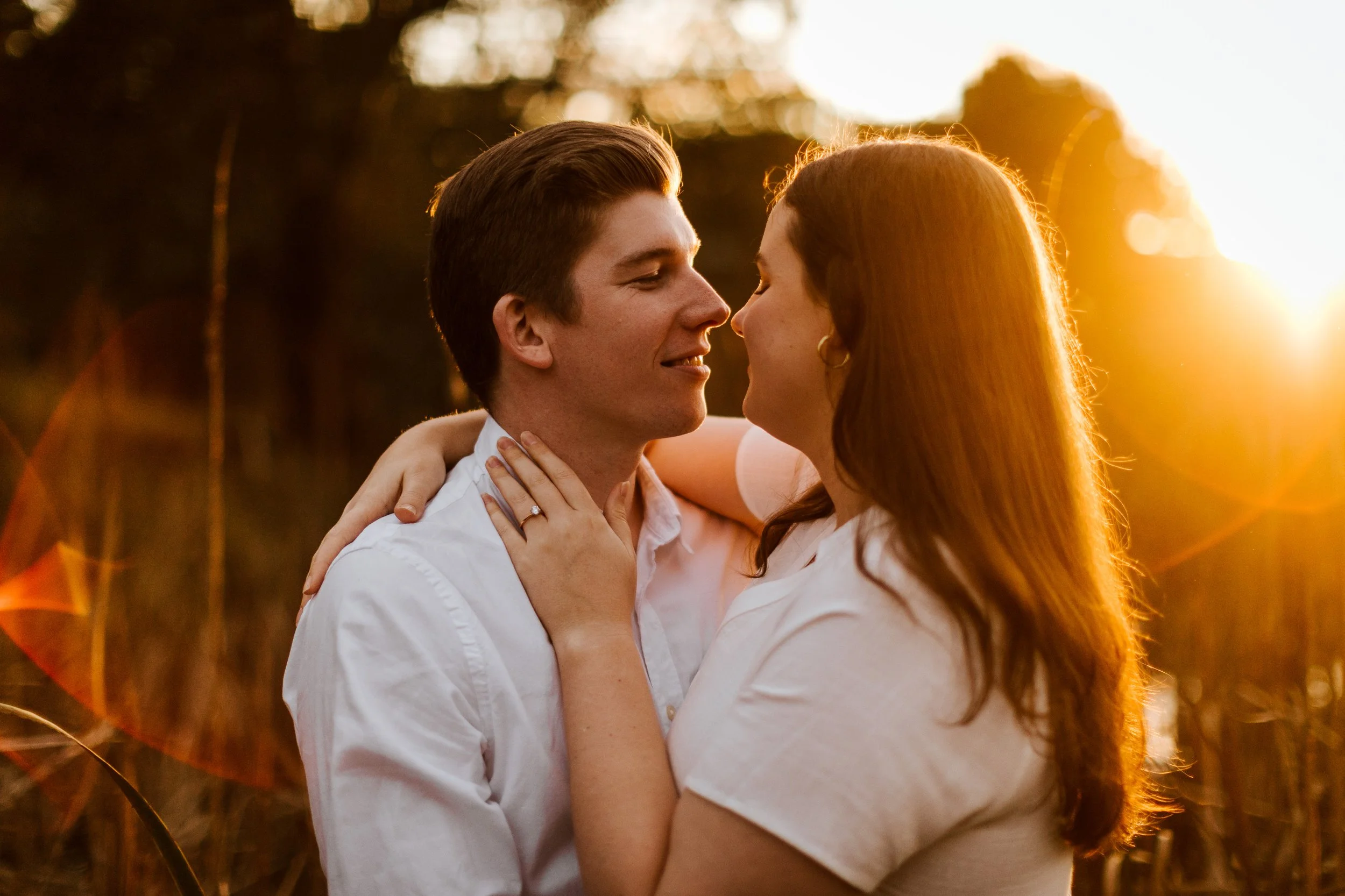 A couple sharing an intimate moment outdoors at sunset, gazing into each other's eyes, with the woman wearing a ring and the man in a white shirt.