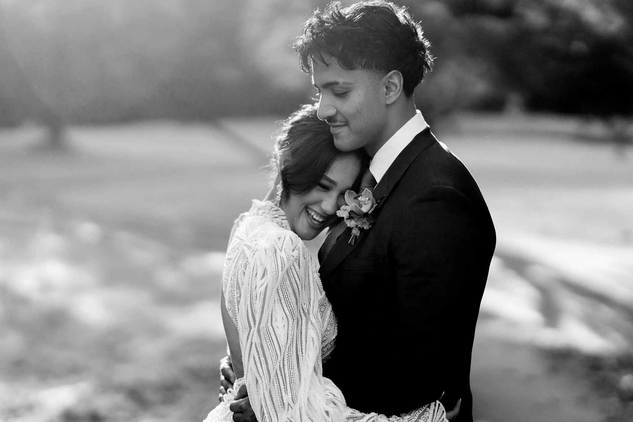 A black and white photograph of a happy couple, embracing each other outdoors, with the woman smiling and resting her head on the man's chest.