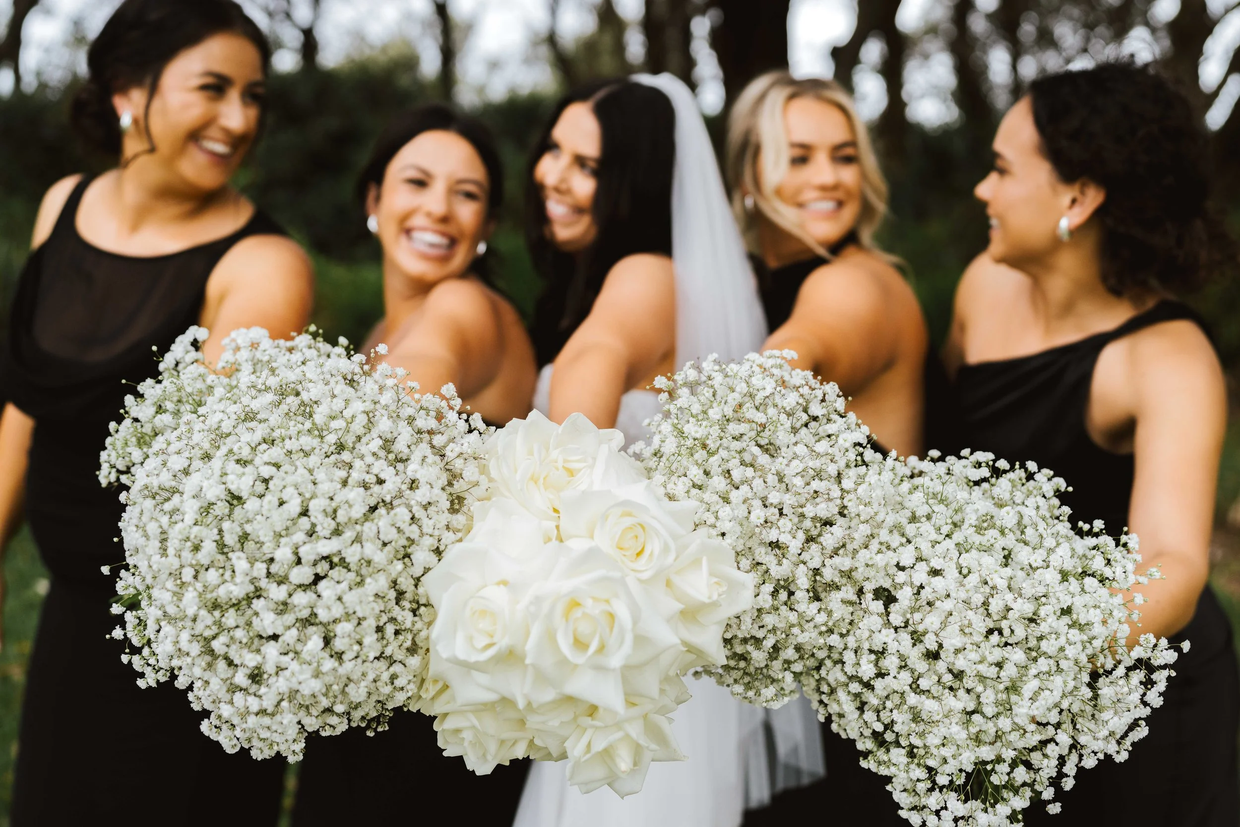 Group of women, including a bride with a veil, smiling and holding a large bouquet of white roses and baby's breath at an outdoor wedding.