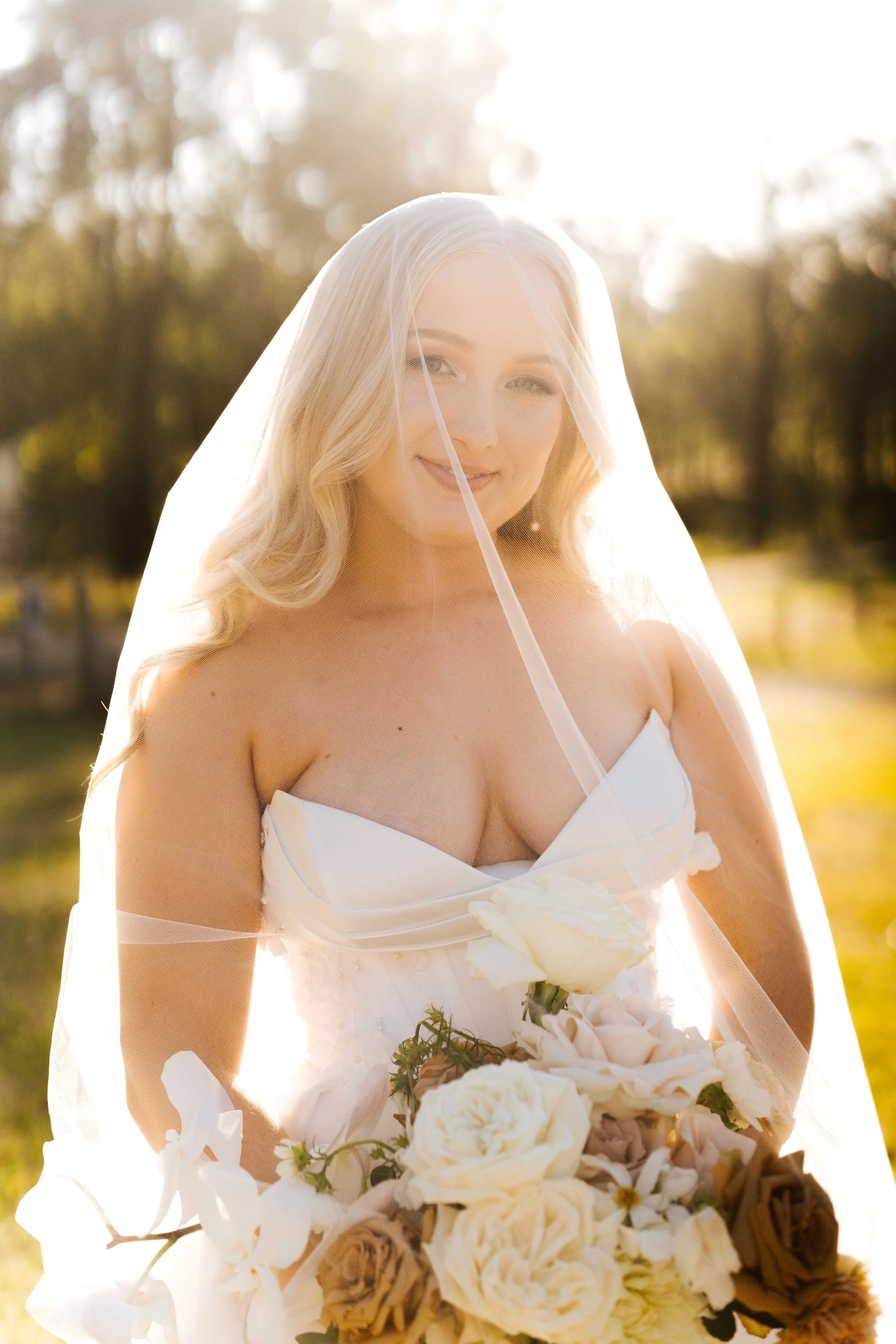 Bride in a white wedding dress holding a bouquet of white and beige flowers, standing outdoors in sunlight with a veil over her face.