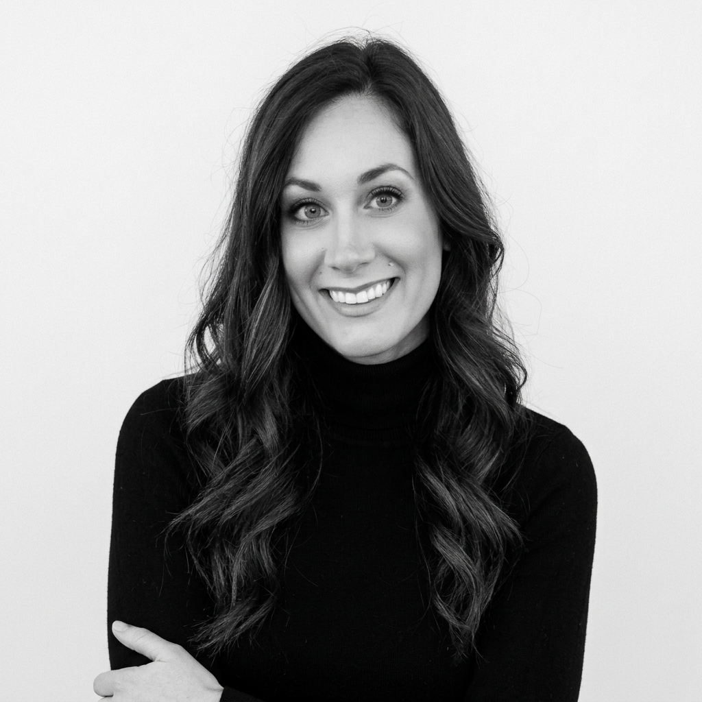 Black and white portrait of a smiling woman with long wavy hair, wearing a black turtleneck sweater, standing against a plain light background.