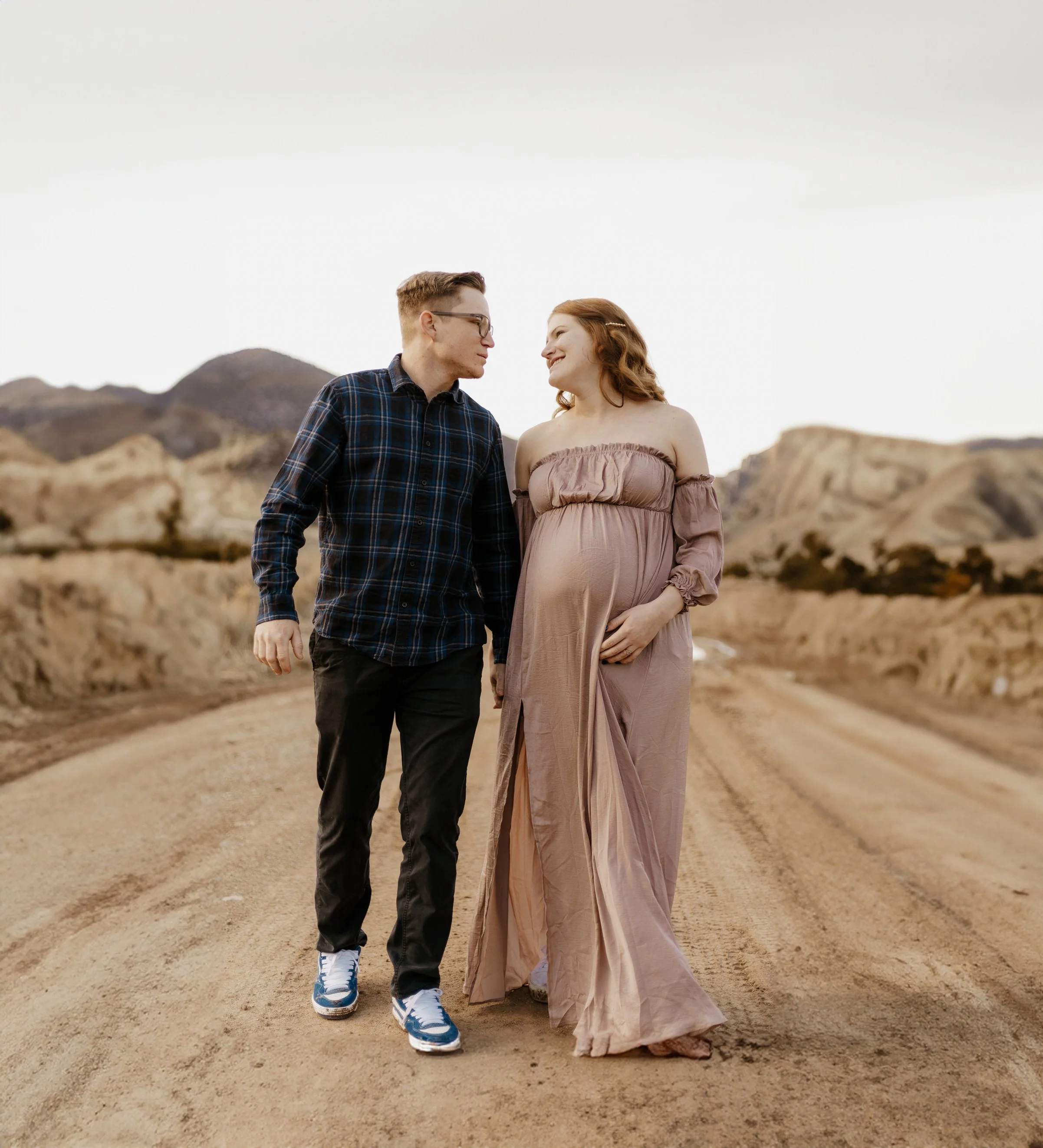 A pregnant woman and a man walking together on a dirt road in a desert landscape, smiling and looking at each other.