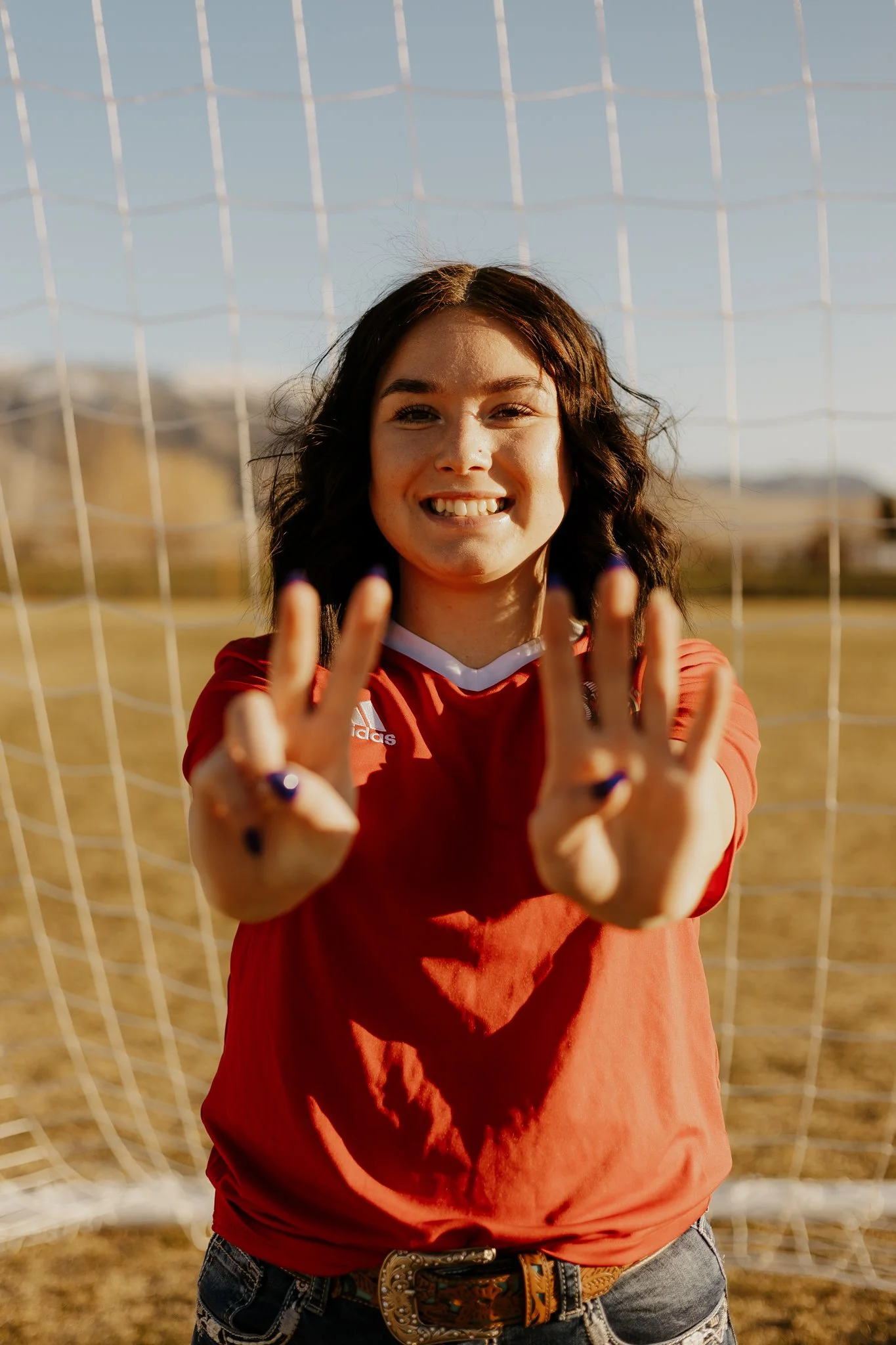 A young woman in a red sports jersey standing in front of a soccer goal, smiling and showing her fingers with dark purple nail polish.