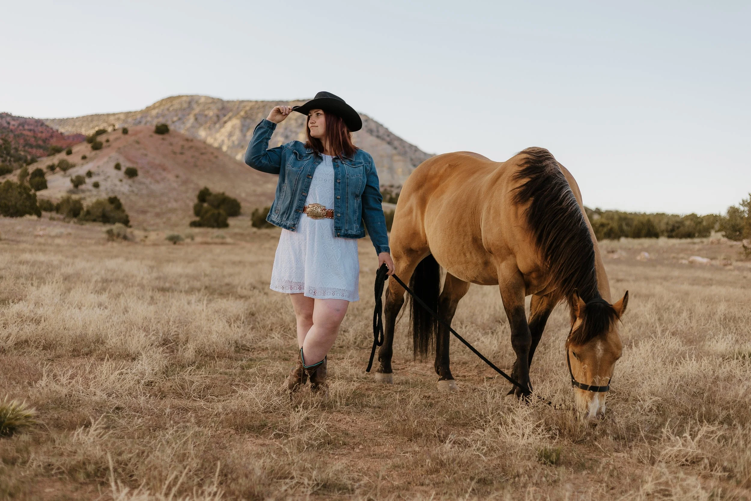 A woman wearing a white dress, denim jacket, cowboy boots, and a black cowboy hat stands in a grassy field holding a leash attached to a tan horse with a black mane. The woman is adjusting her hat while looking into the distance. In the background, there are mountains and a clear sky.
