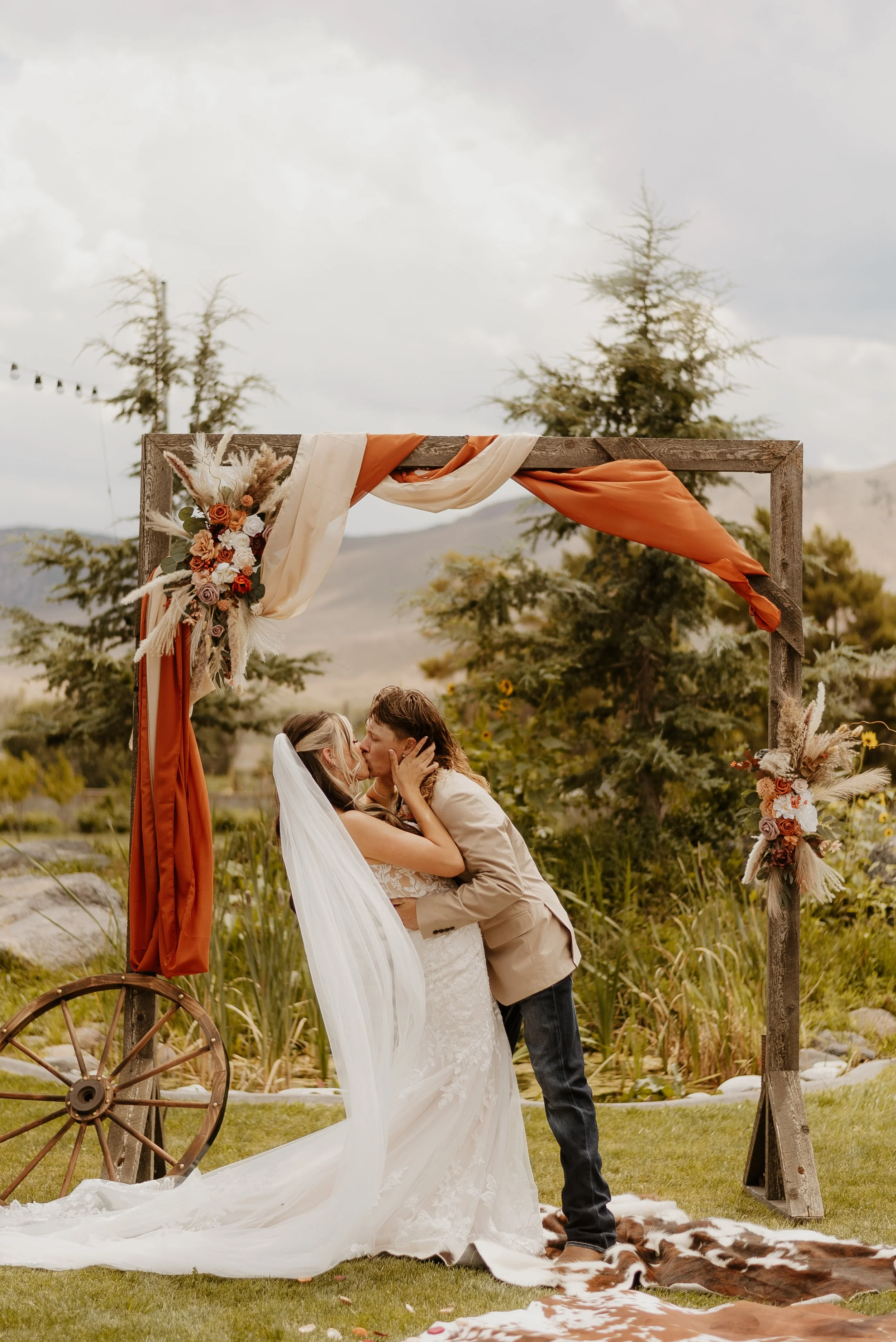 A couple in wedding attire sharing a kiss under a decorated wooden arch outside, with greenery and mountains in the background.