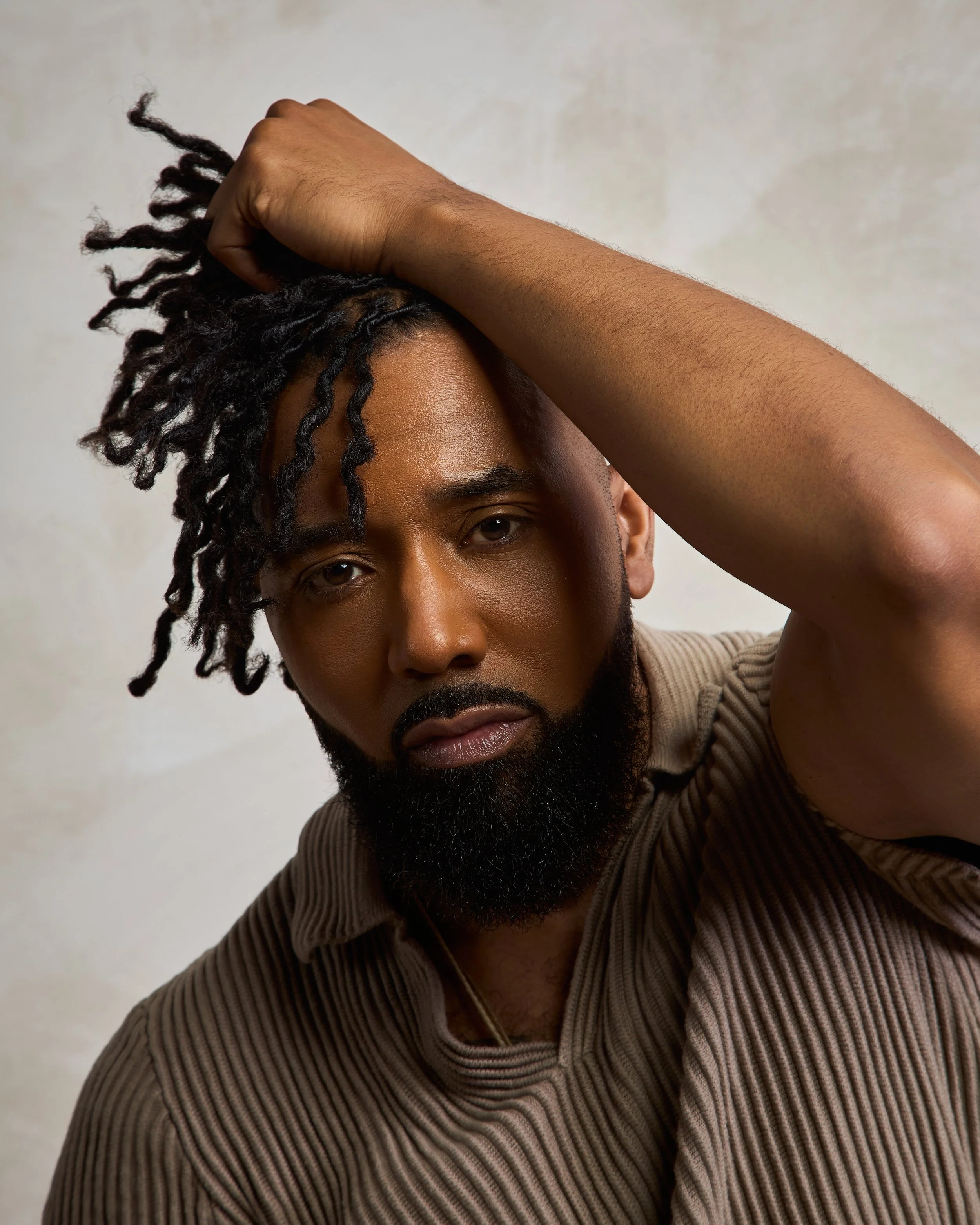 A man with a beard and dreadlocks looking directly at the camera with his arm raised, resting on his head, wearing a beige ribbed shirt against a neutral background.