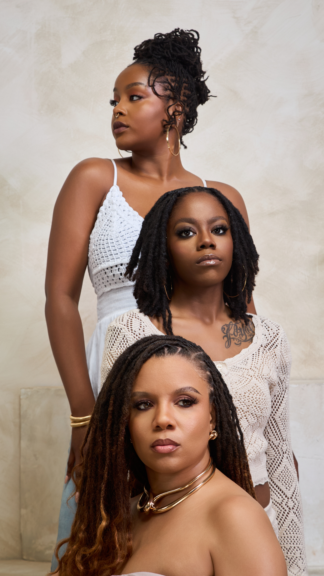 Three women with dark skin and dreadlocked hair posing together against a beige background. Each woman is dressed in light-colored, stylish clothing and wearing jewelry, with confident expressions.