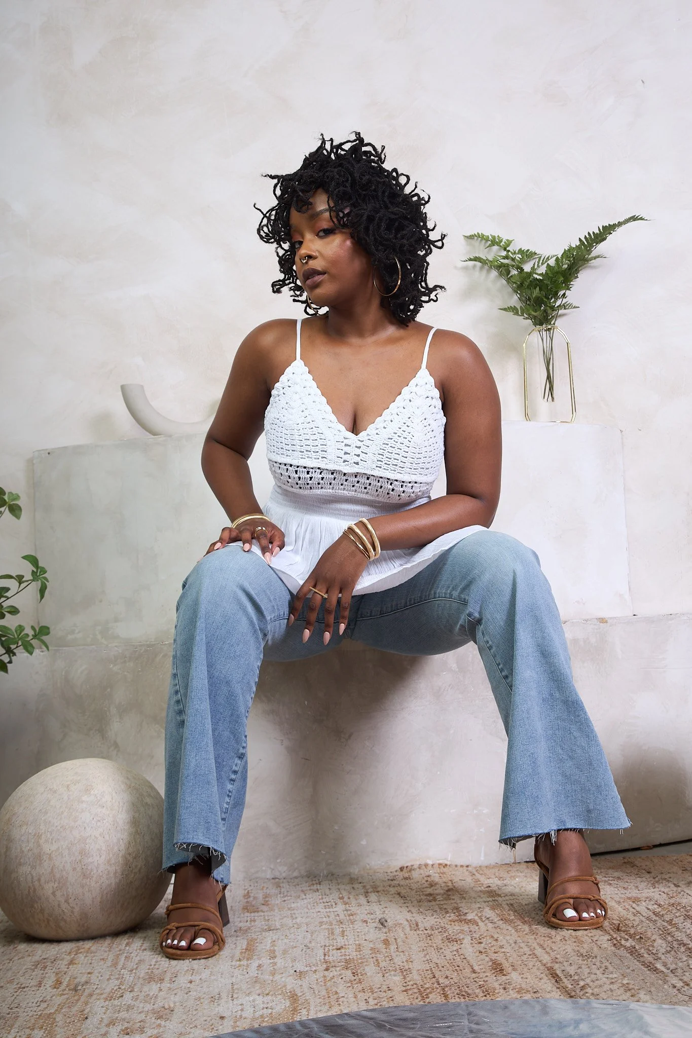 A woman with dark, curly hair wearing a white crochet tank top, light blue jeans, and tan heeled sandals, sitting on a ledge with her hands on her knees and looking slightly to the side. There is a beige ball on the floor, a small plant on the left, 