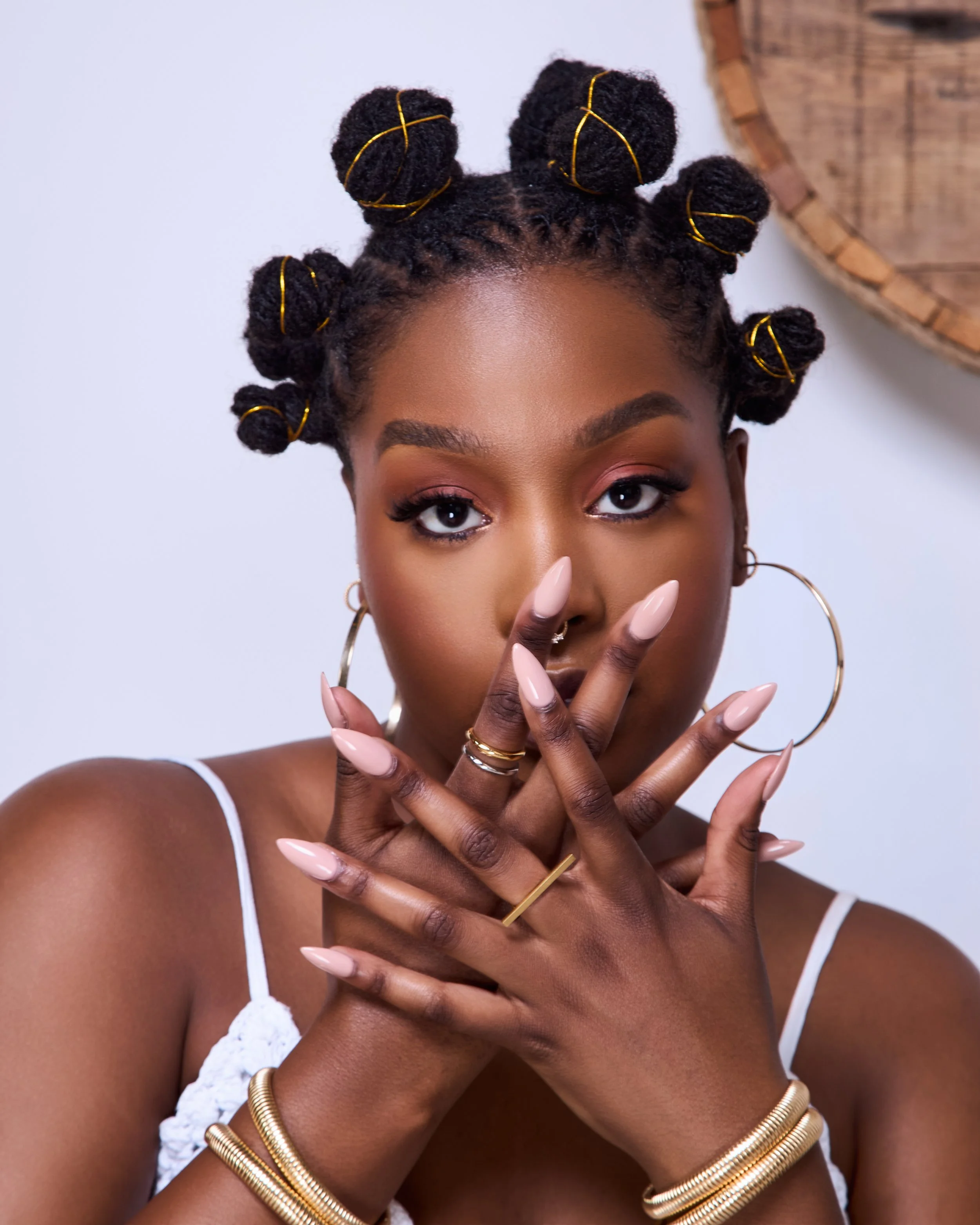 Close-up of a woman with styled hair wrapped in yellow wire, wearing large hoop earrings, rings, and gold bracelets, with a white tank top against a plain background.