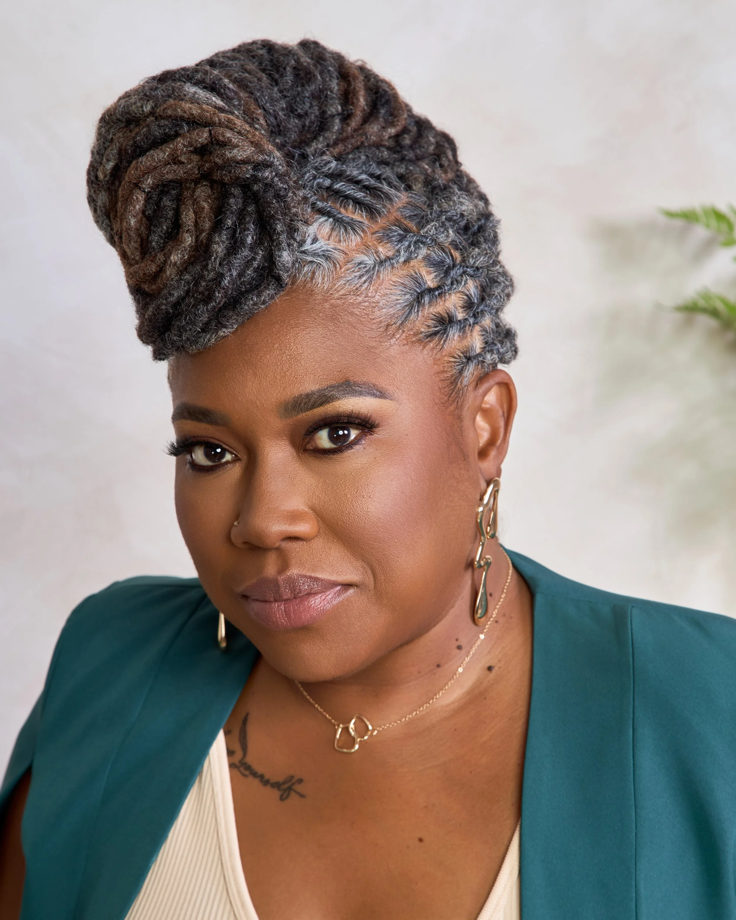 A woman with dark skin and styled dreadlocks in an updo, wearing a green blazer, gold earrings, and a necklace, posed in front of a neutral background with a plant visible on the right.