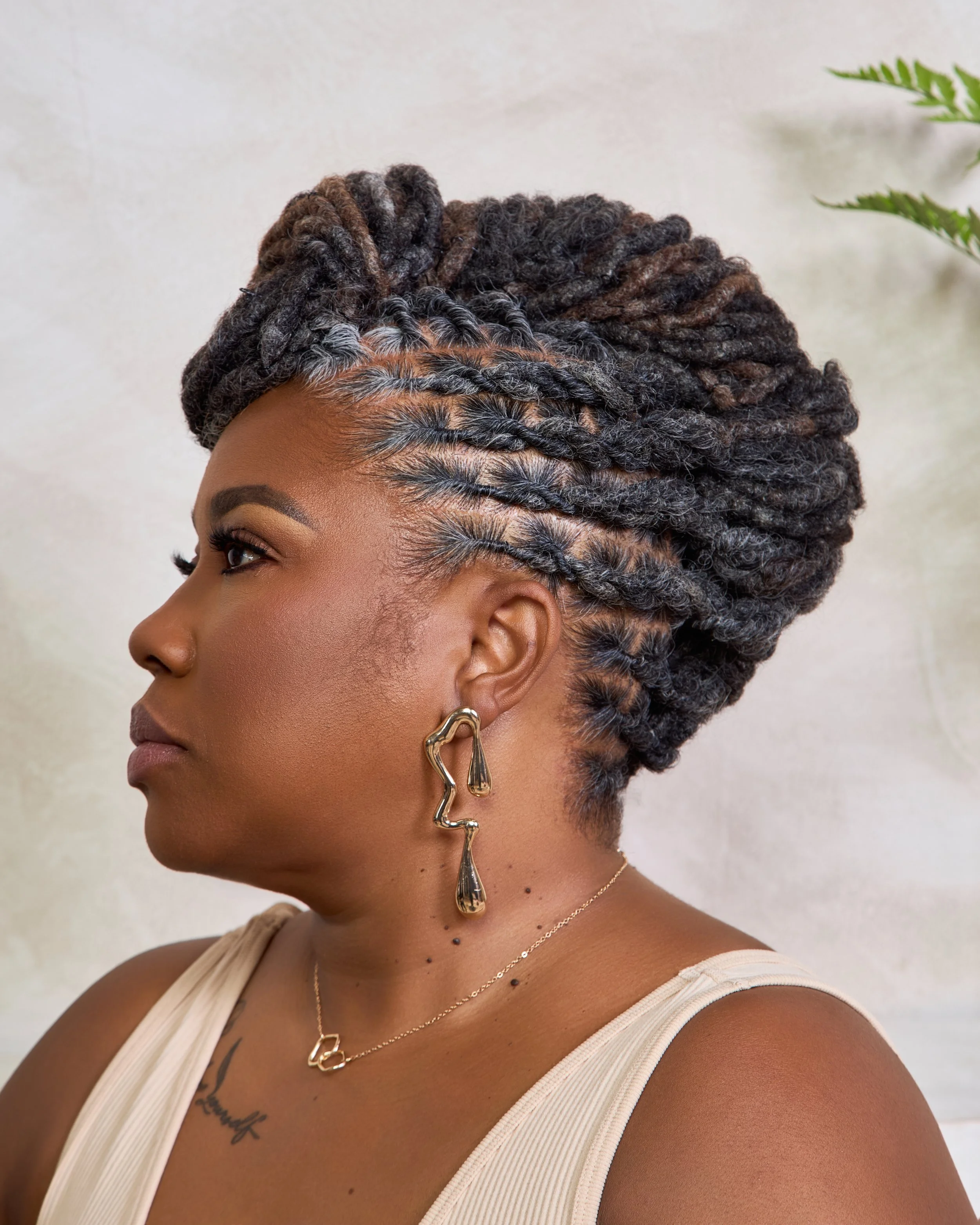 A woman with styled dreadlocks in a twisted updo, wearing a beige sleeveless top, gold earrings, a necklace, and with a tattoo on her chest, standing in front of a neutral background and some green leaves.