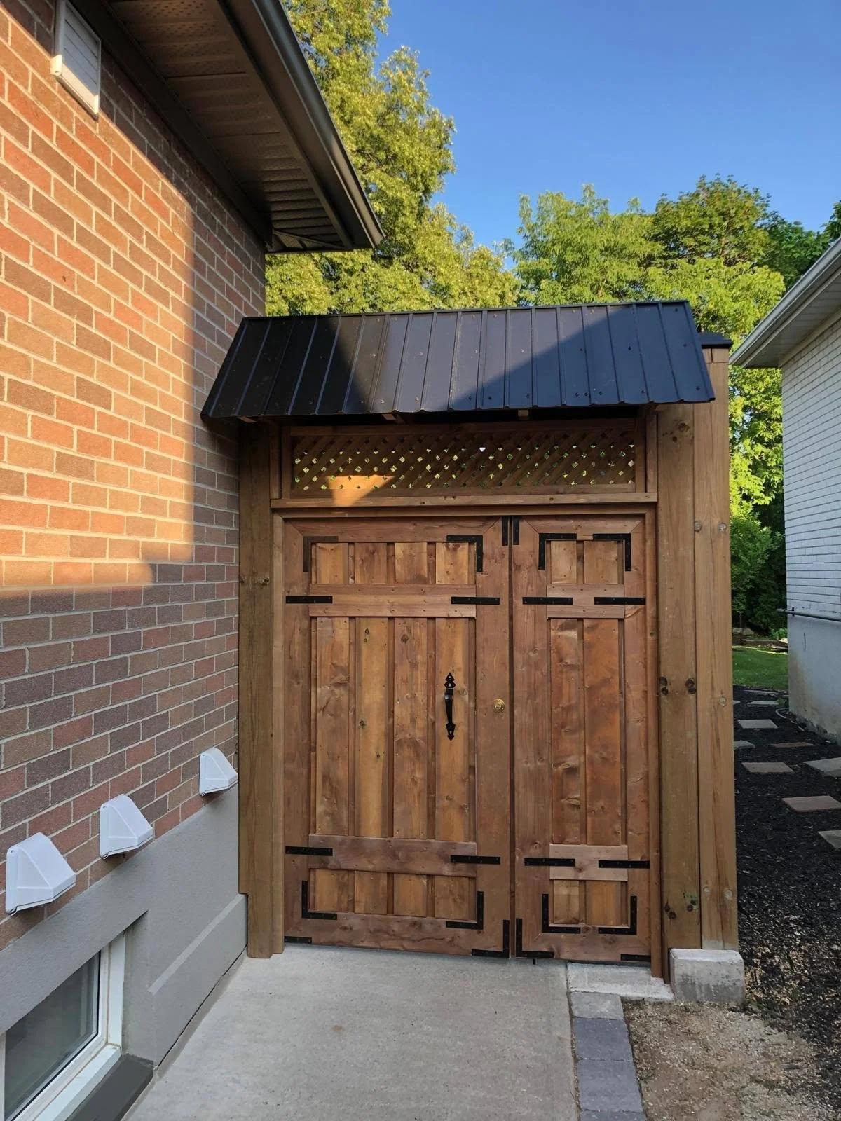 Outdoor wooden storage enclosure with double doors and black metal hinges beside a brick house.