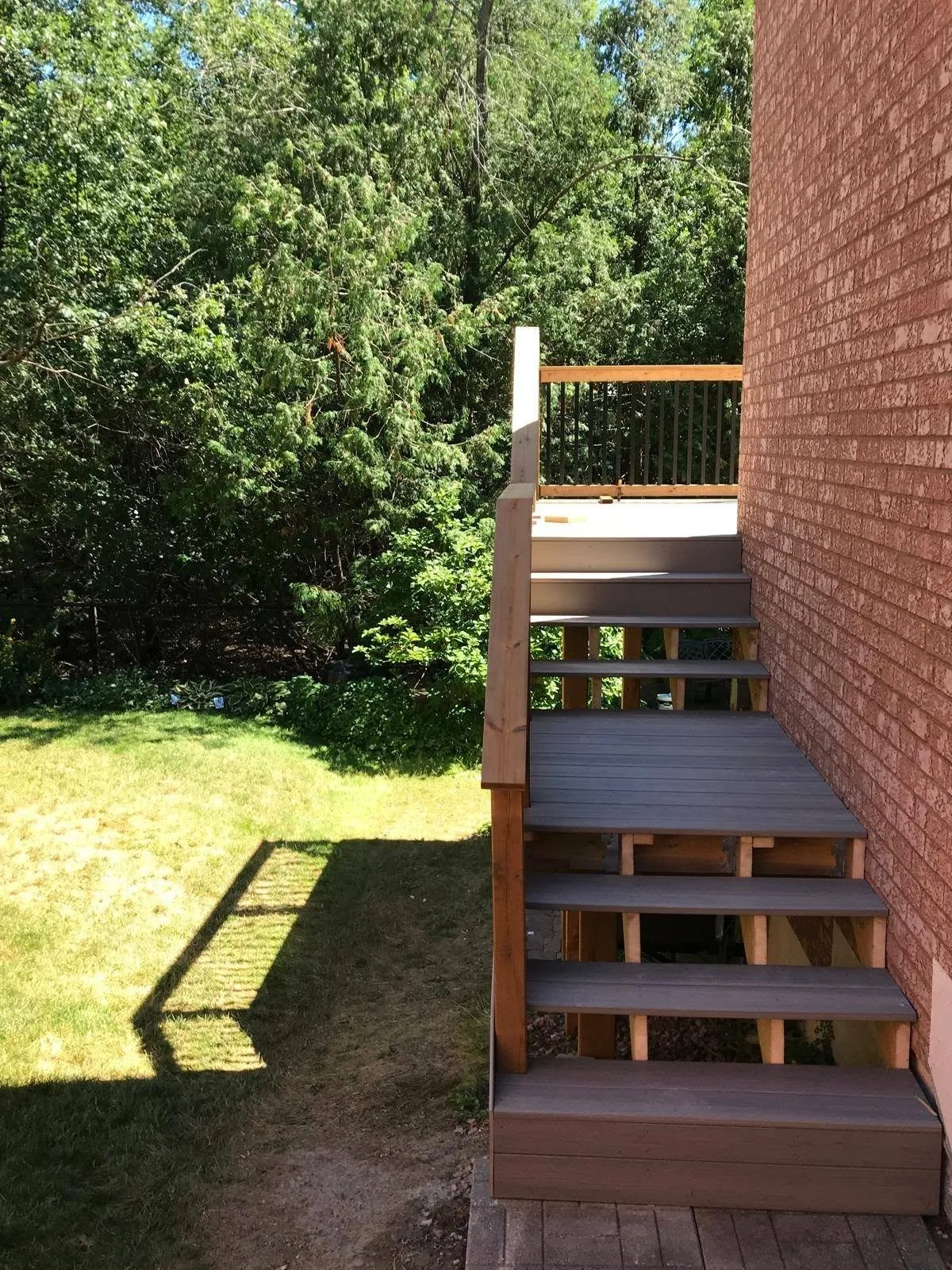 Outdoor wooden deck staircase with brown timber steps and railings leading to an elevated deck beside a brick house.
