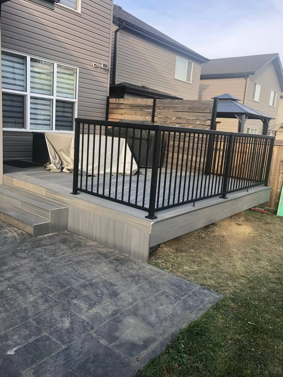 Raised backyard deck with black metal railings and a decorative stamped concrete patio beside a residential house.