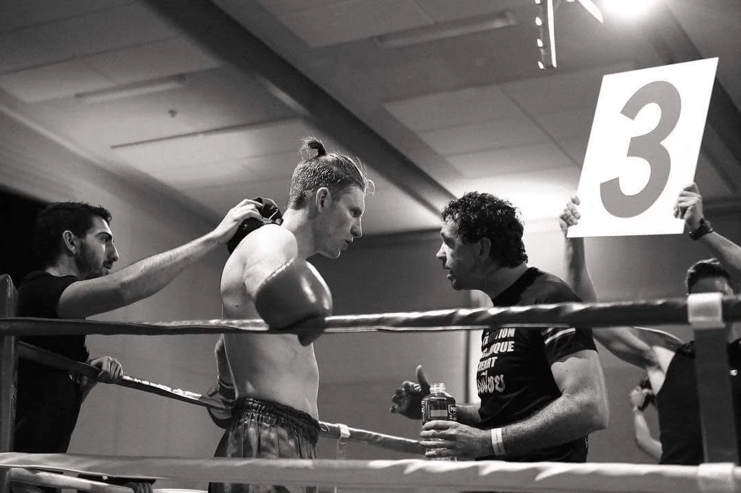 Boxer and coach having a serious pre-fight conversation inside a boxing ring, with a person holding a sign with the number 3.