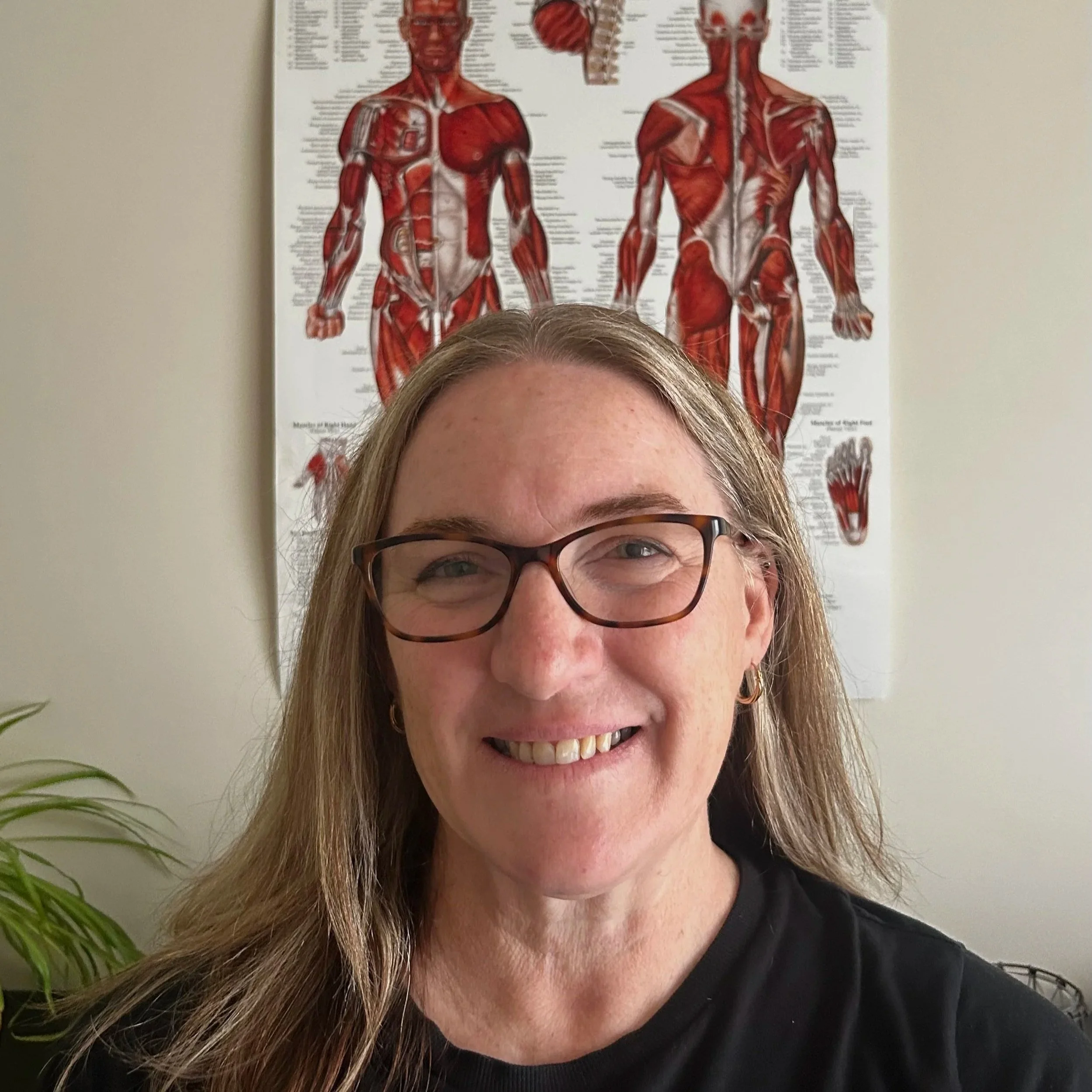 A smiling woman with glasses and long blonde hair taking a selfie in front of a human anatomy poster on a beige wall. The poster shows detailed illustrations of muscular anatomy.