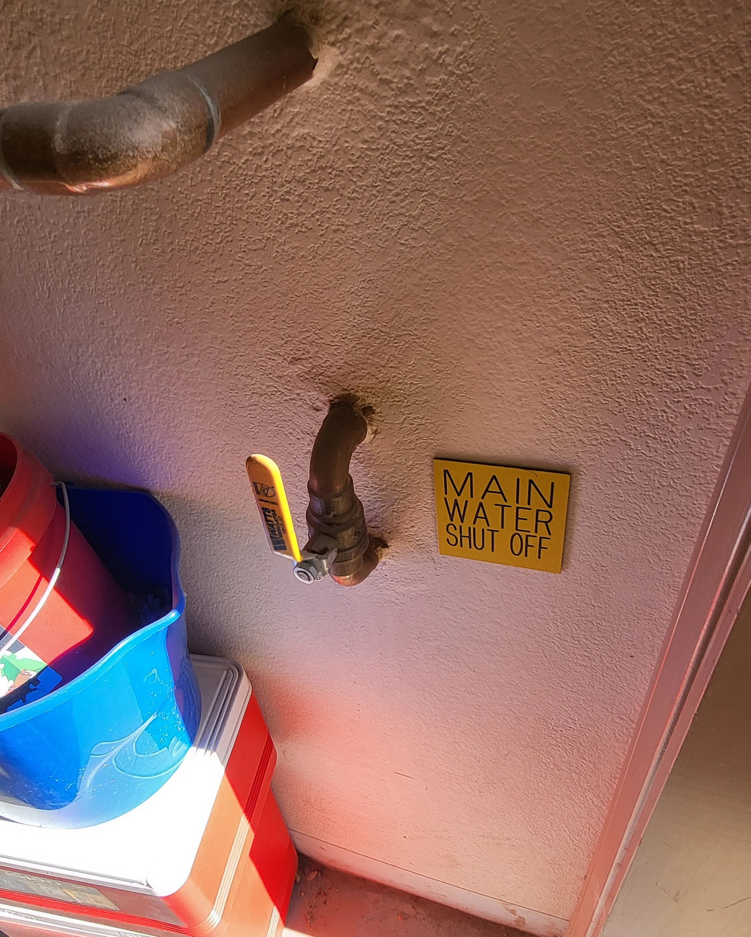 A water faucet with a yellow handle and a sign that says 'Main Water Shut Off' on a textured wall, with a blue container and a red cooler in the corner.
