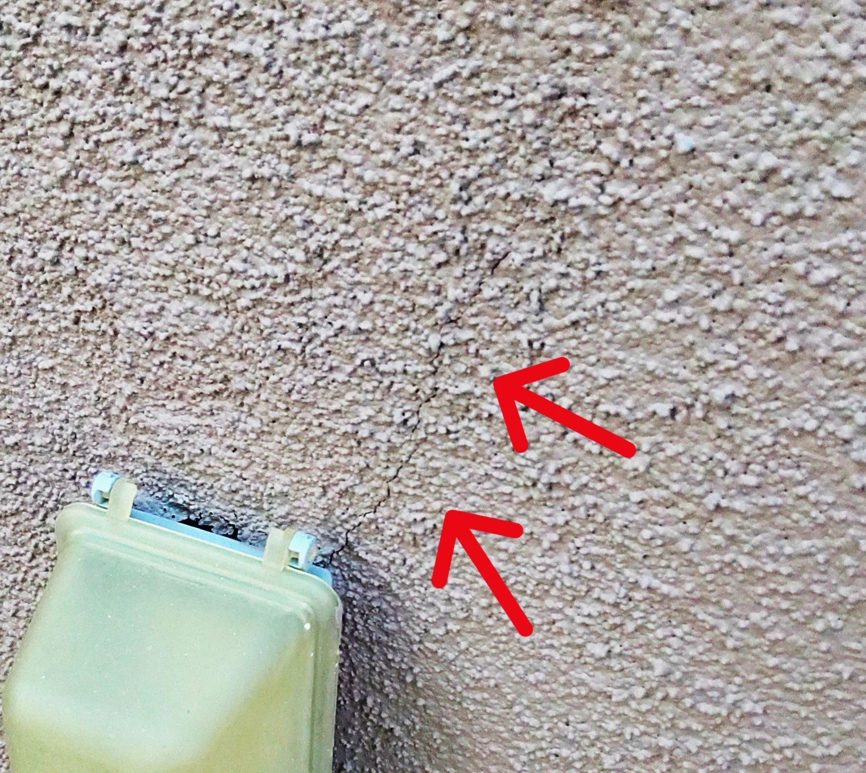 Close-up photo of a textured beige wall with two red arrows pointing to a visible crack running vertically.