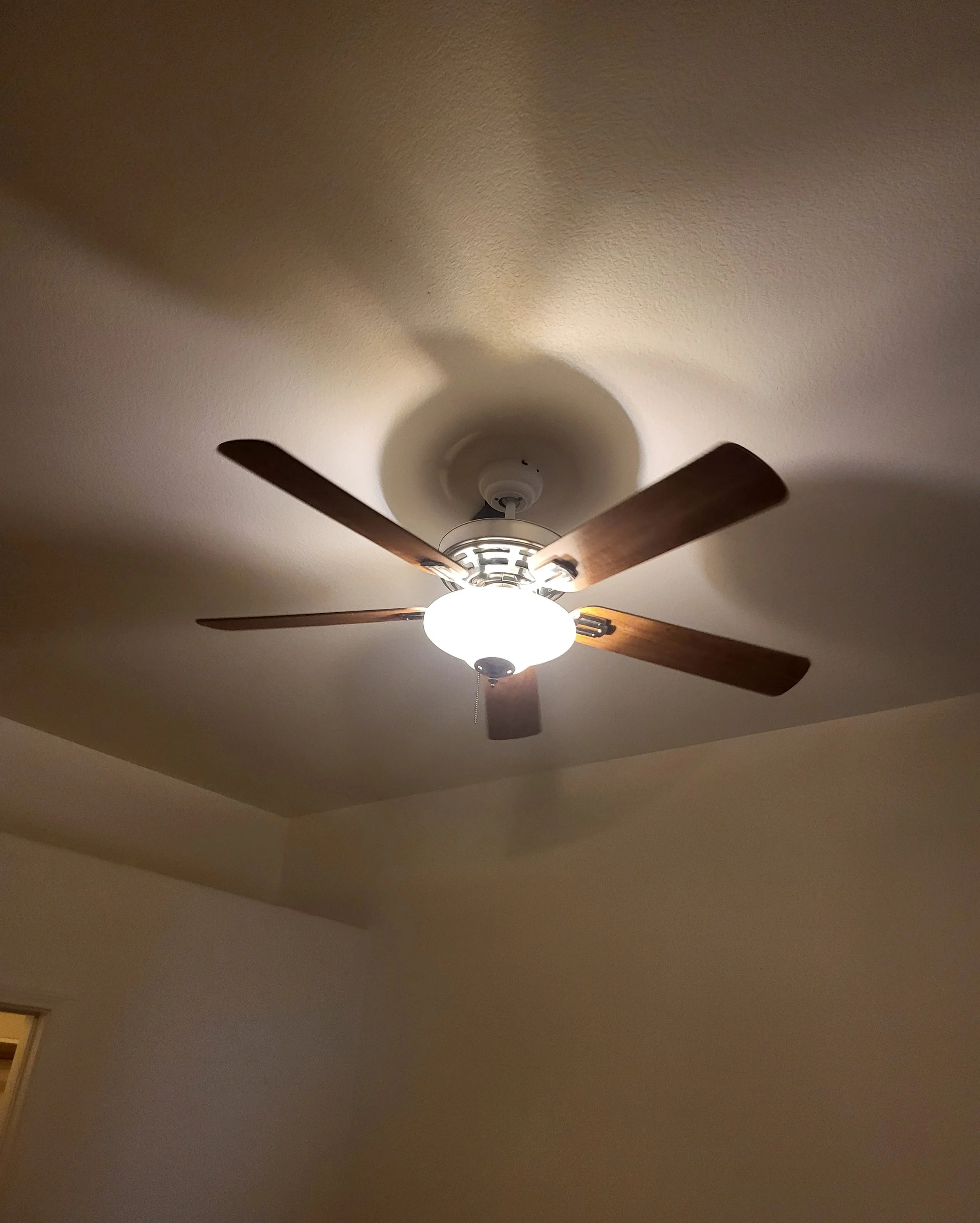 Ceiling fan with lights turned on, mounted on a beige ceiling in a corner of a room.