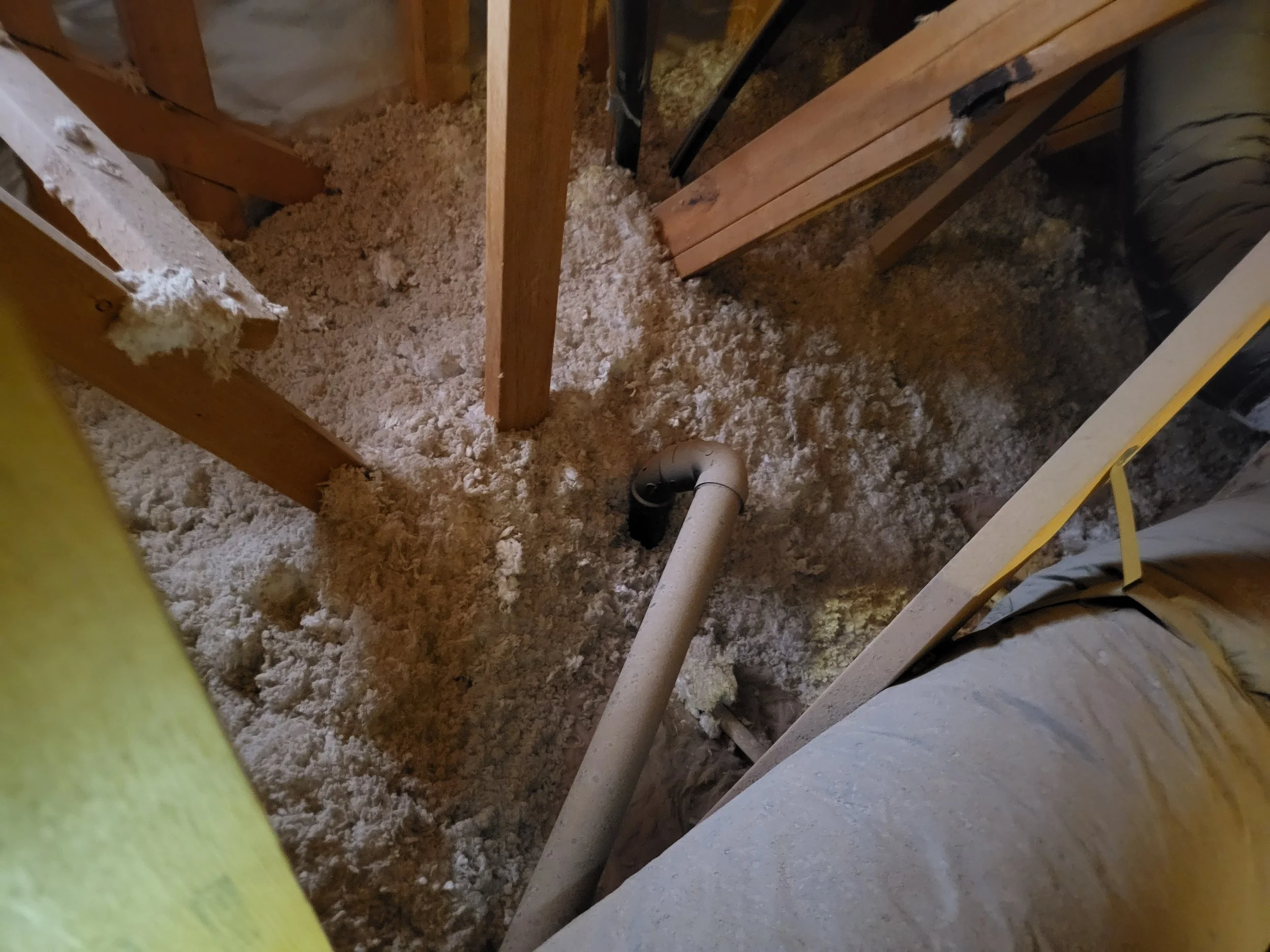 Insulation and plumbing pipes in an unfinished attic space with exposed insulation and insulated HVAC Ducting.