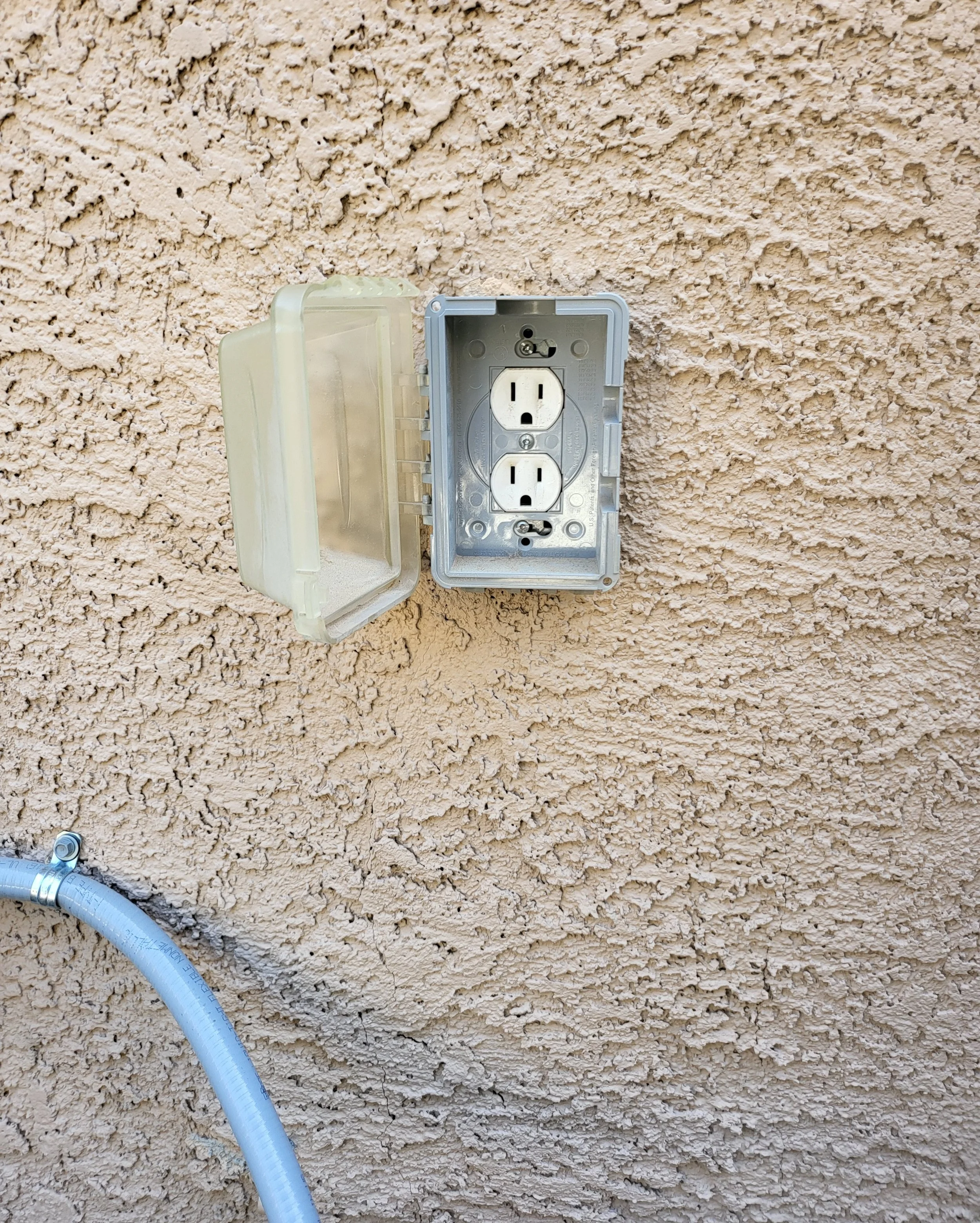 An outdoor weatherproof electrical outlet with a cover, mounted on a textured beige wall. A grey conduit is visible at the bottom left corner.