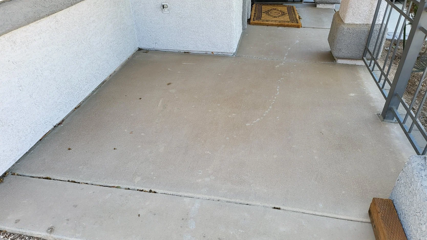 Front porch with a concrete floor, white stucco walls, and a metal railing on the right. A small doormat is near the door, and a wooden block is at the bottom right corner.