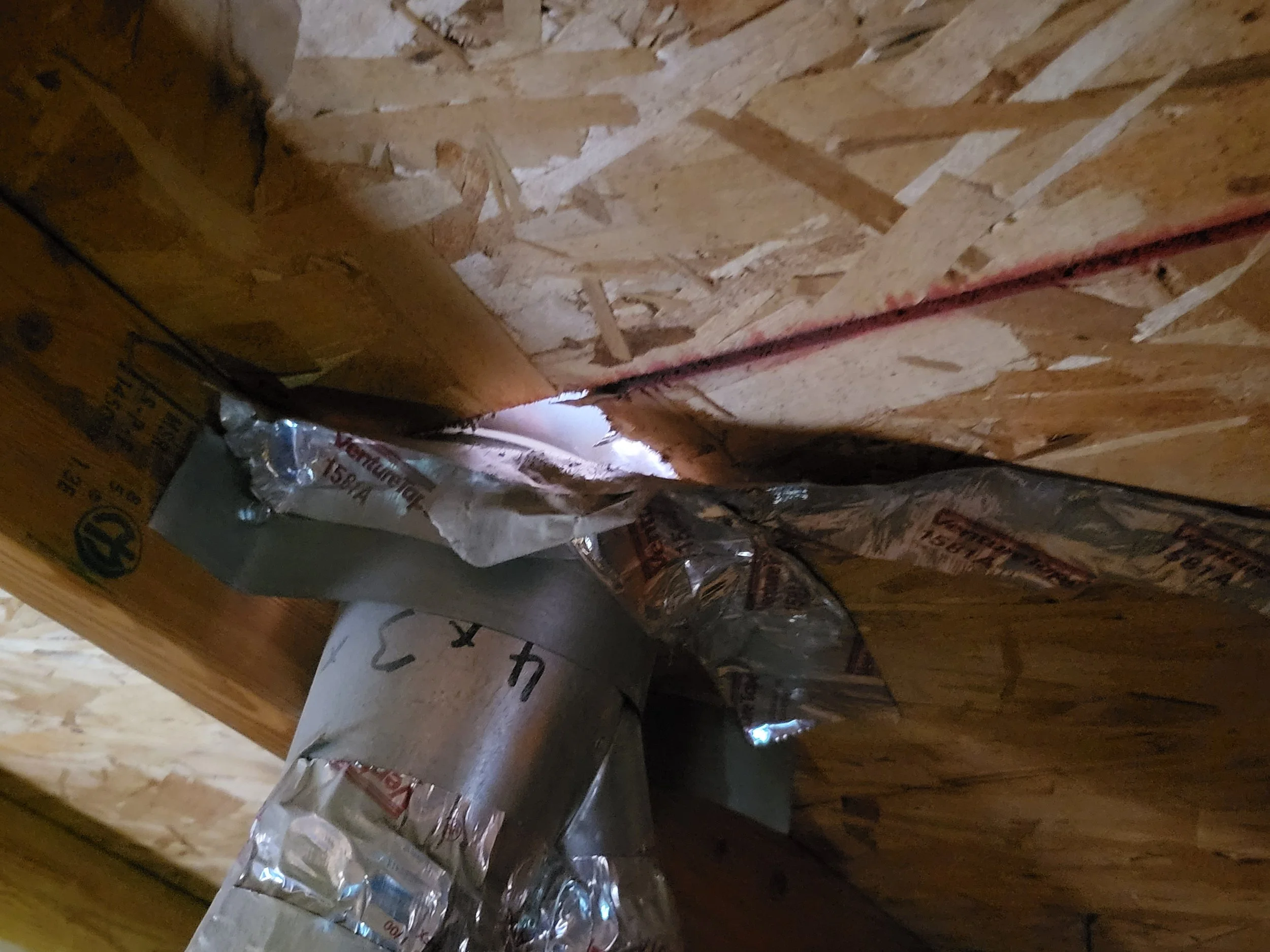 View of a metal pipe passing through an opening in a wooden wall with insulation and foil tape around the pipe.