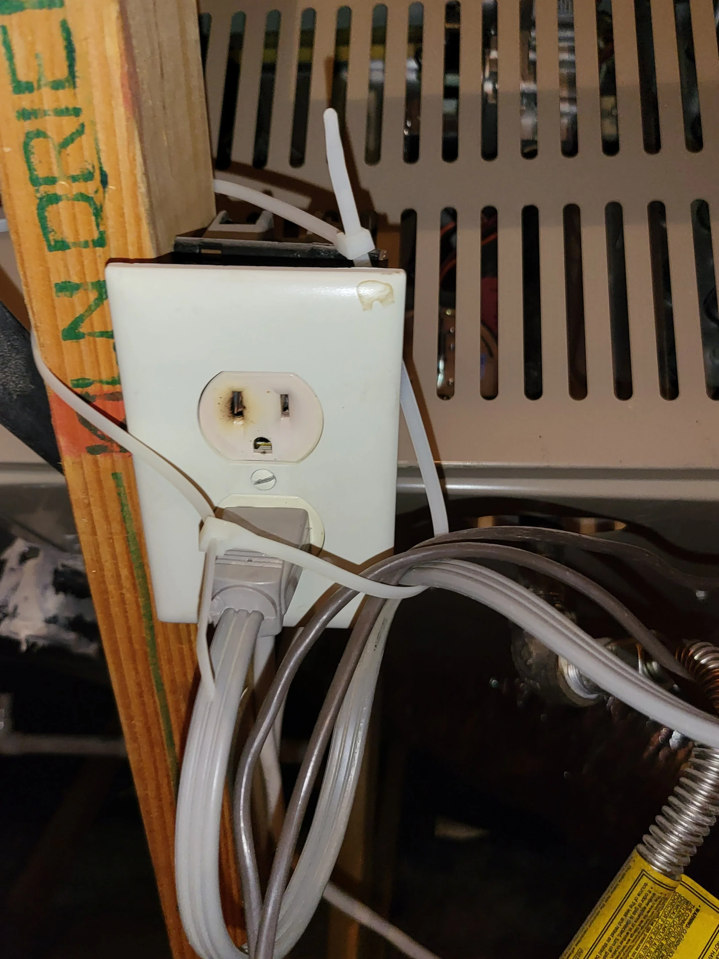 Electrical outlet with multiple cords plugged in, mounted on a wooden surface next to a metallic structure.