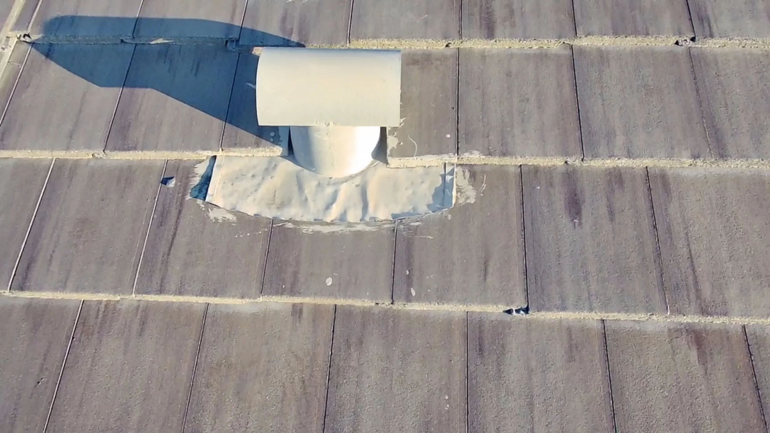 Close-up of a rooftop vent pipe with a weatherproof cover, installed on a brown shingle roof, casting a shadow.