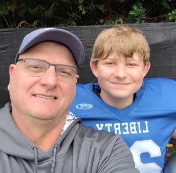 A man wearing glasses, a cap, and a gray hoodie taking a selfie with a young boy in a blue sports jersey, both smiling outdoors with a black fence and green plants in the background.