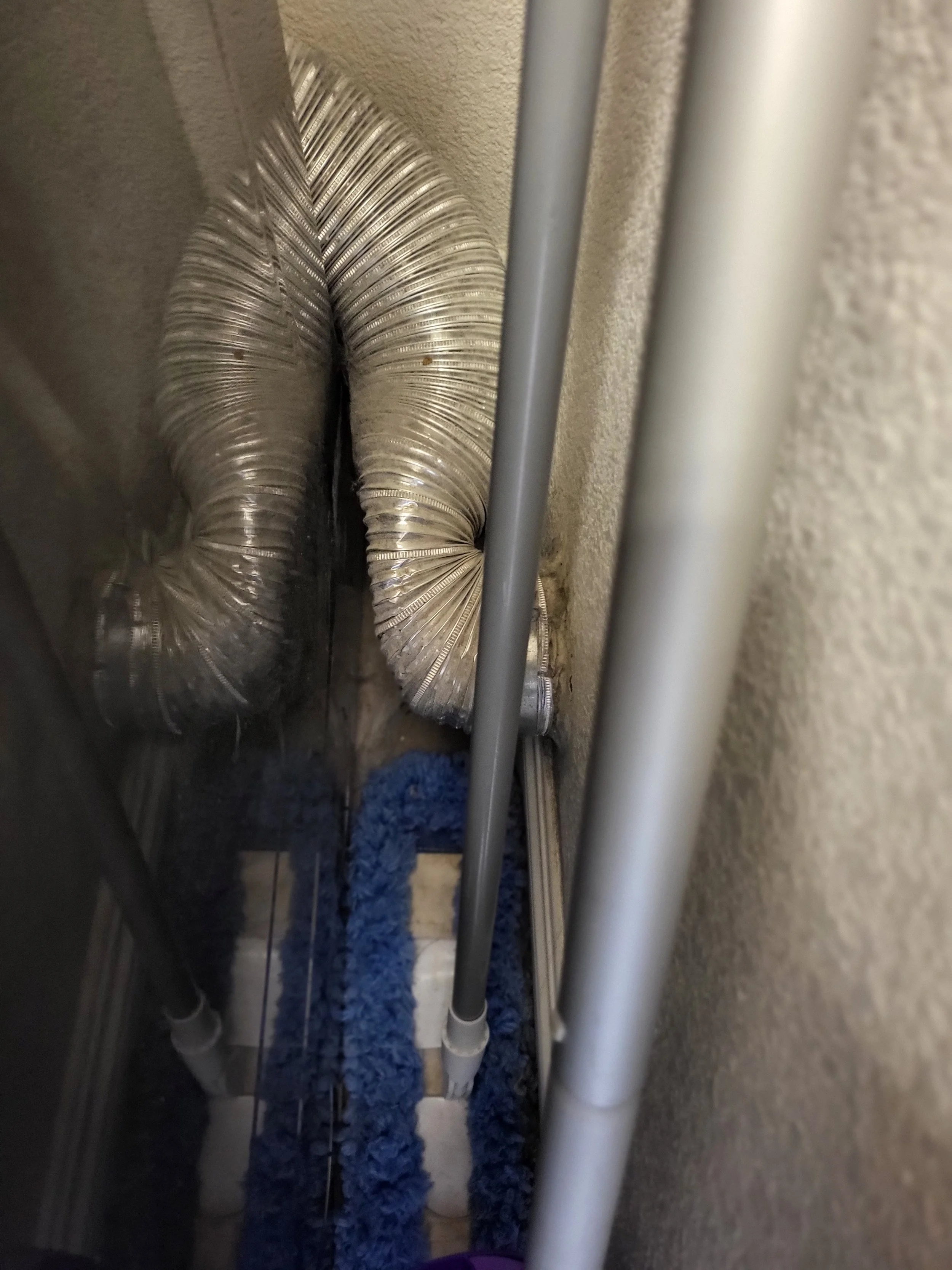 Close-up view of silver flexible air ducts, gray and white pipes, and a blue cleaning mop or dusting tool in a narrow space behind a wall.