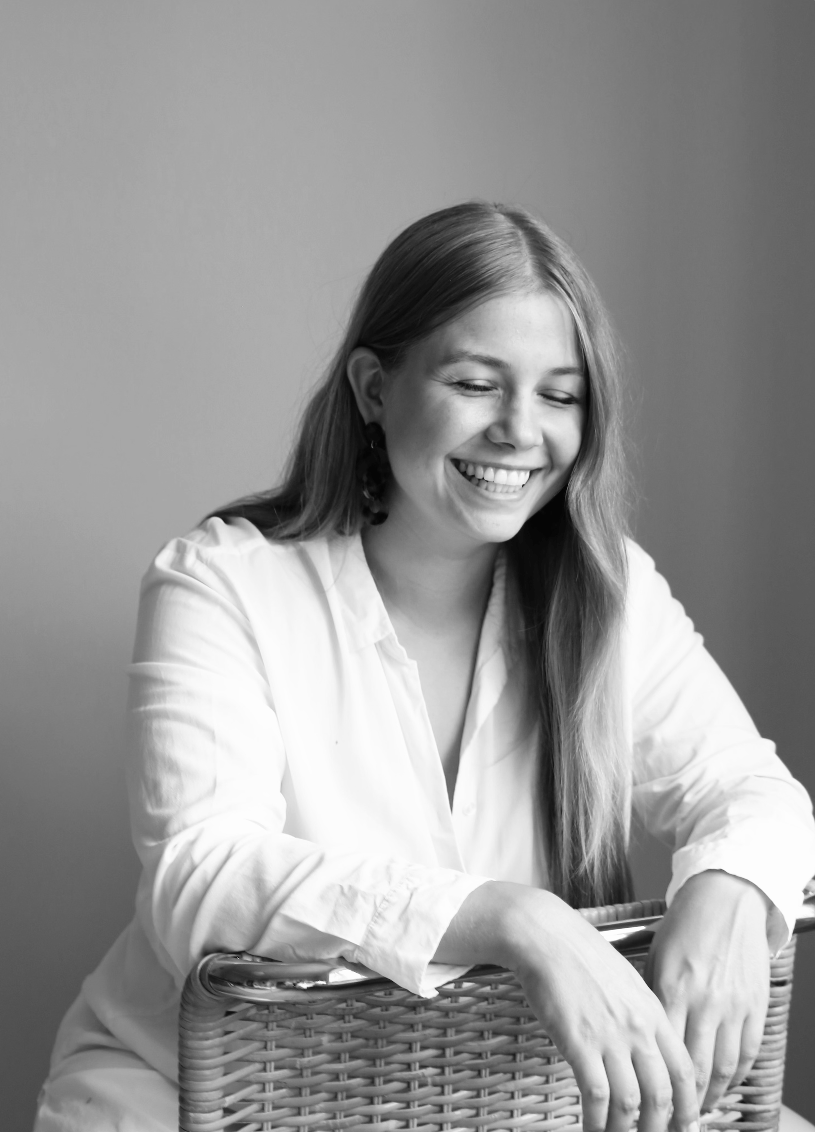 Black and white photo of a young woman with long hair smiling, sitting in a wicker basket, wearing a white shirt, against a plain gray background.