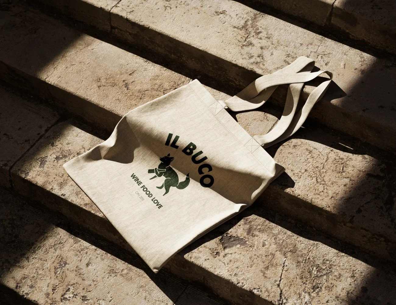 A beige tote bag with the text ‘Il Buco’ and a cartoon fox illustration, lying on stone stairs with sunlight casting shadows.