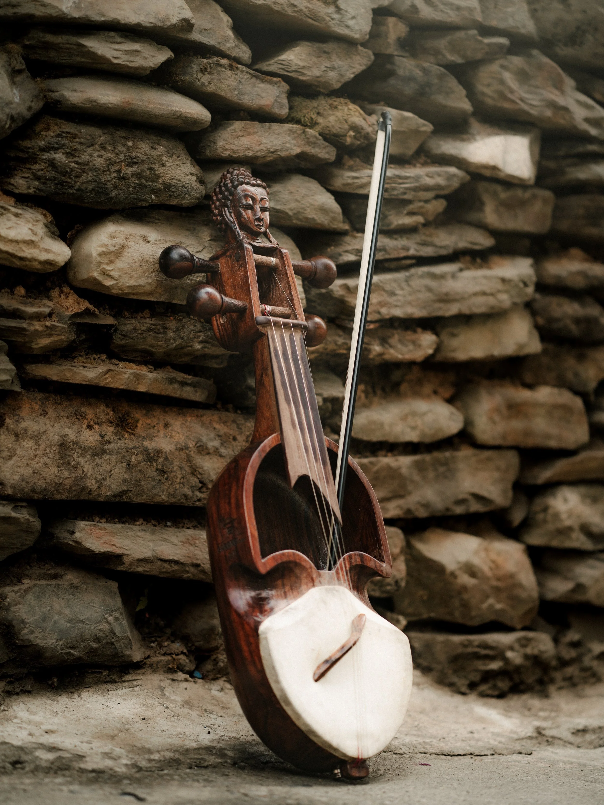 A wooden theremin resembling a sarangi with a carved buddha head on top, set against a stone wall background.