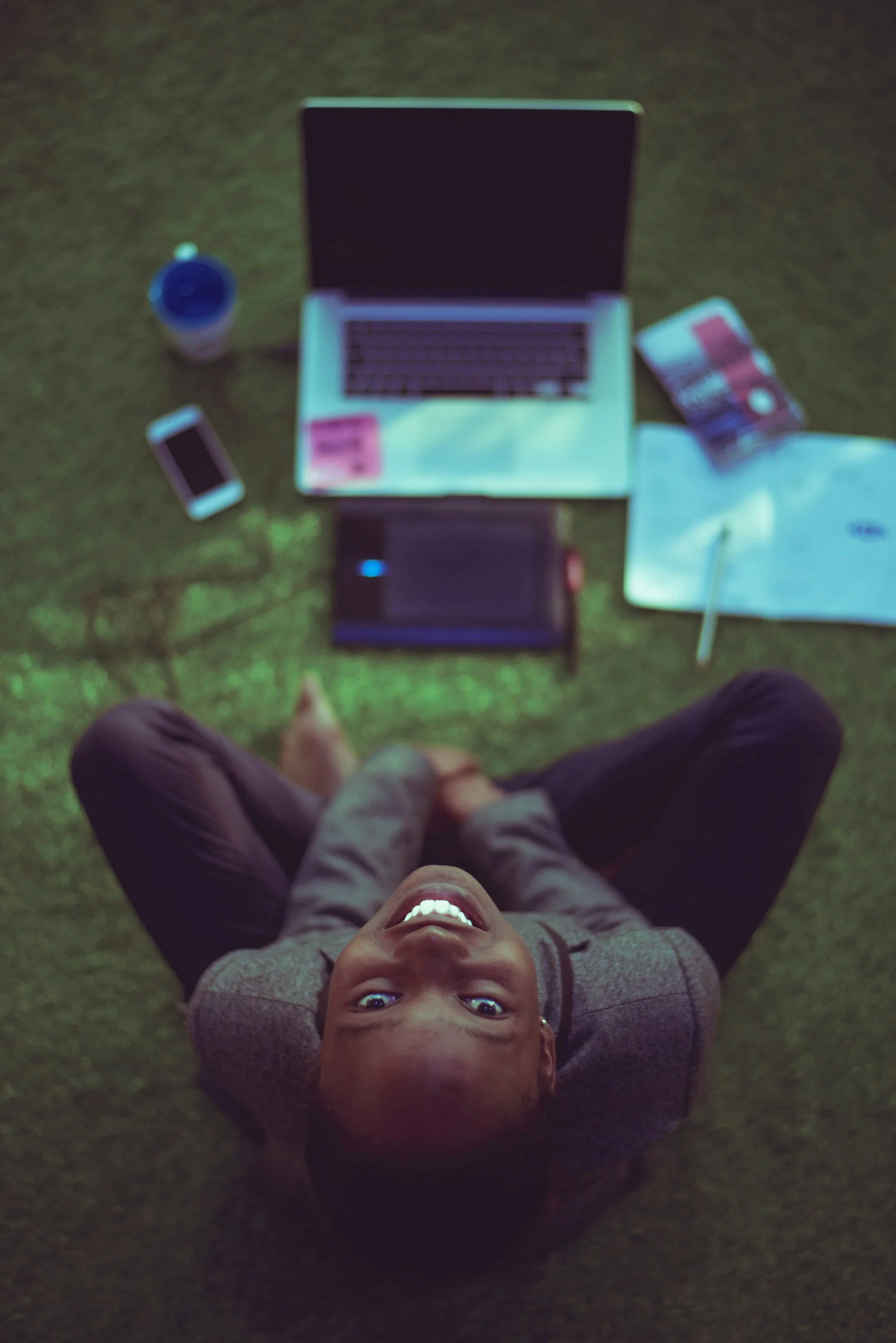 A young boy lying on the grass looking up at the camera with a big smile, surrounded by a laptop, smartphone, cup, snacks, and a notebook.