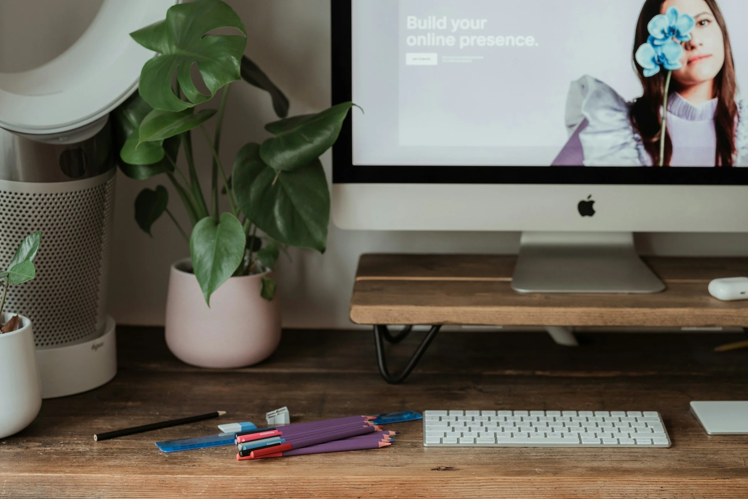 A workspace with a desktop computer, a potted Monstera plant, colored pens, and a keyboard on a wooden desk. In the background, a monitor displays a website with the text 'Build your online presence' and a woman holding a blue flower.