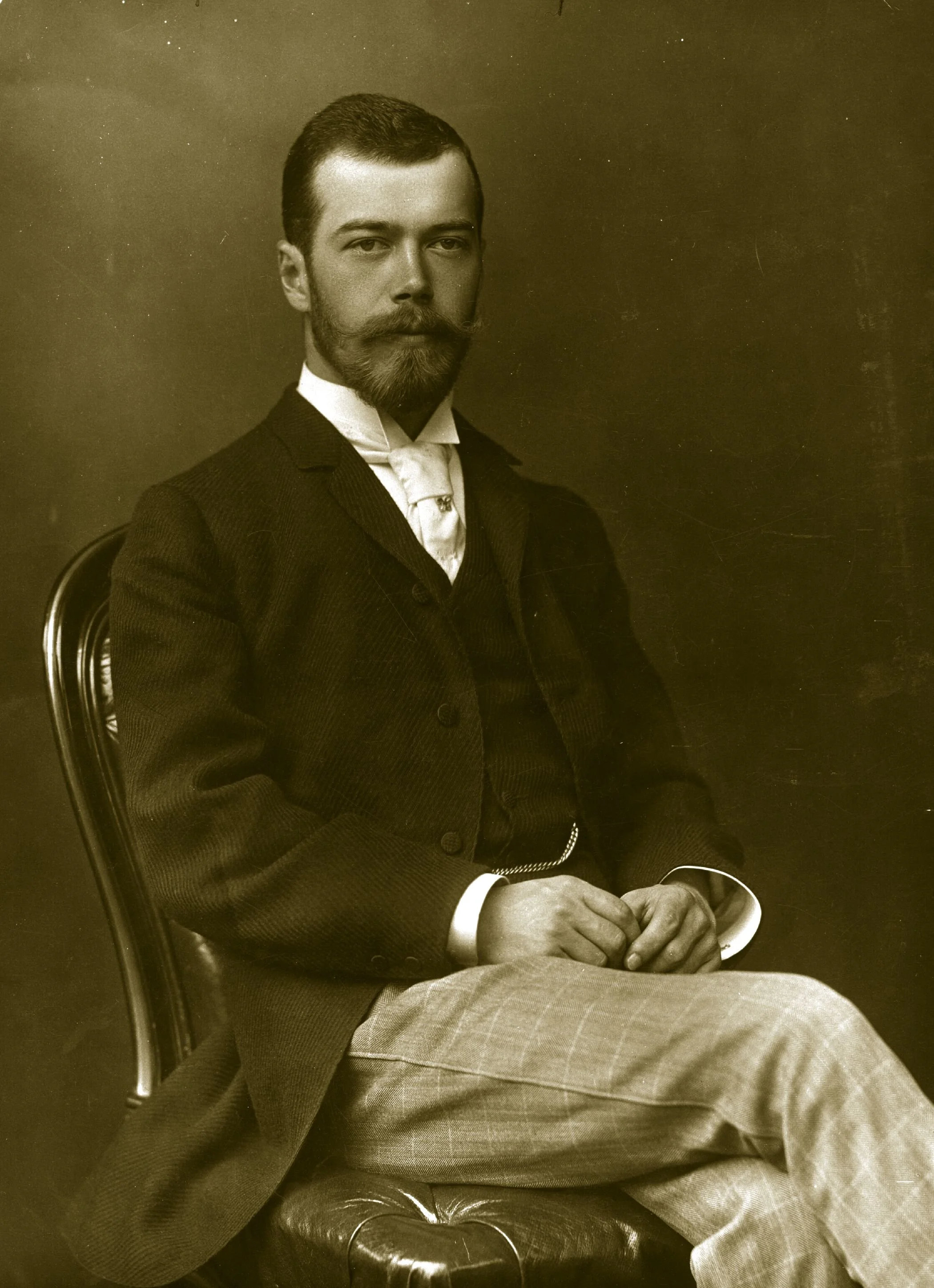 A black-and-white portrait of a man with a beard and mustache, dressed in formal 19th-century attire, sitting on a chair with his hands resting on a cup, against a plain dark background.