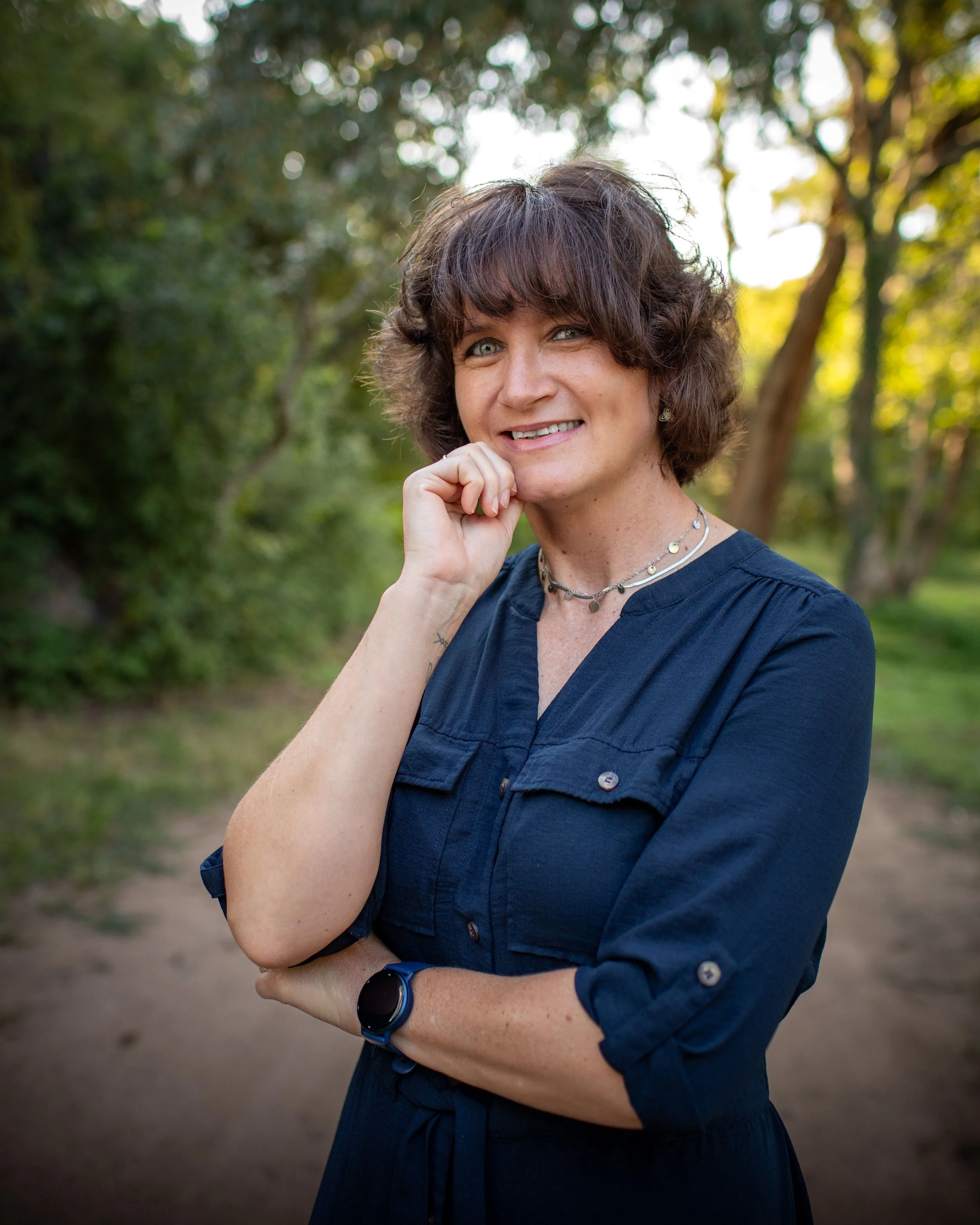 A woman with short curly brown hair and blue eyes standing outdoors in a green, wooded area. She is smiling, with her right hand touching her chin, and wearing a navy blue dress.