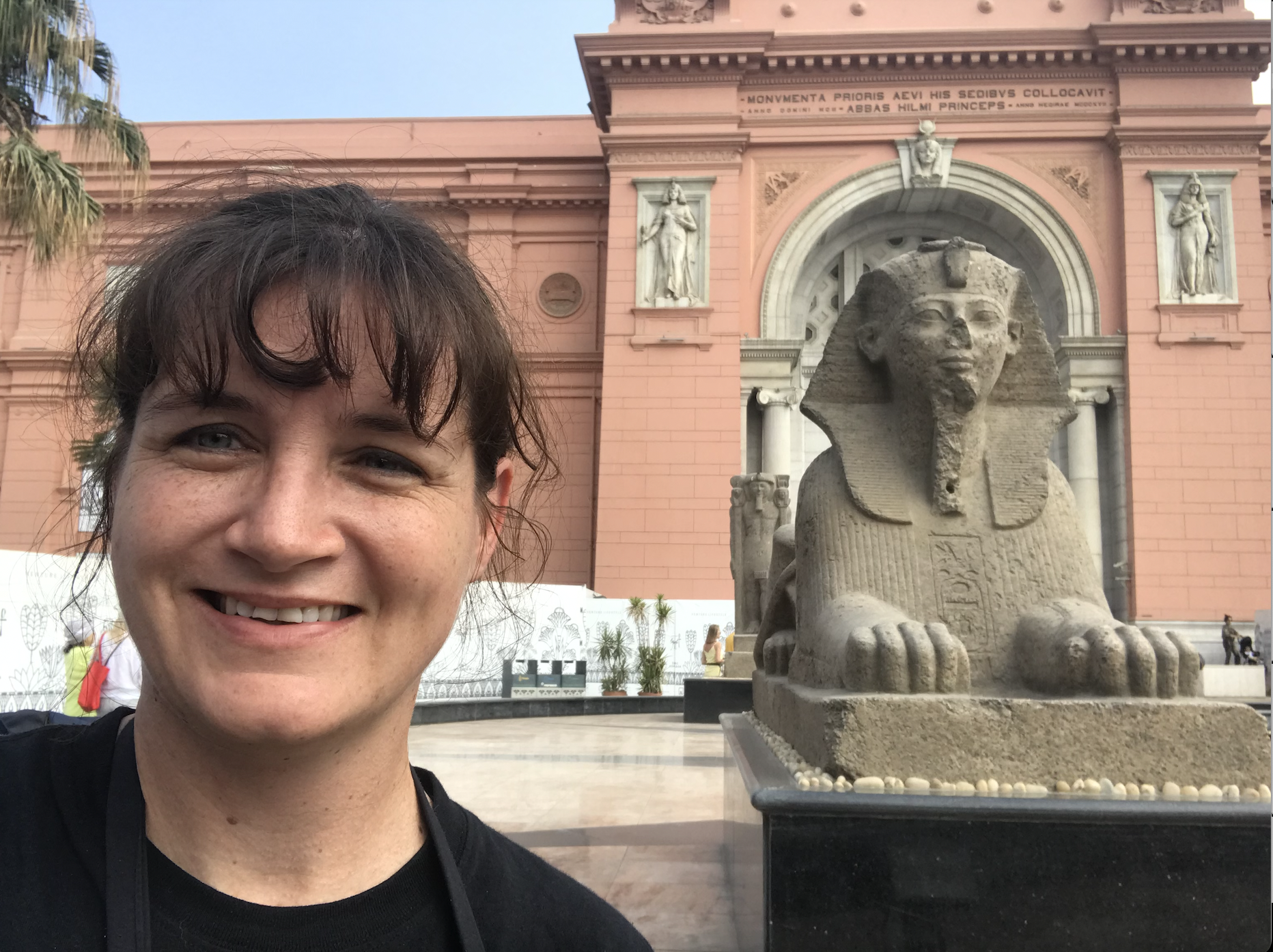 Close-up of a smiling woman with curly brown hair standing in front of an ancient Egyptian Sphinx statue at a historical site.