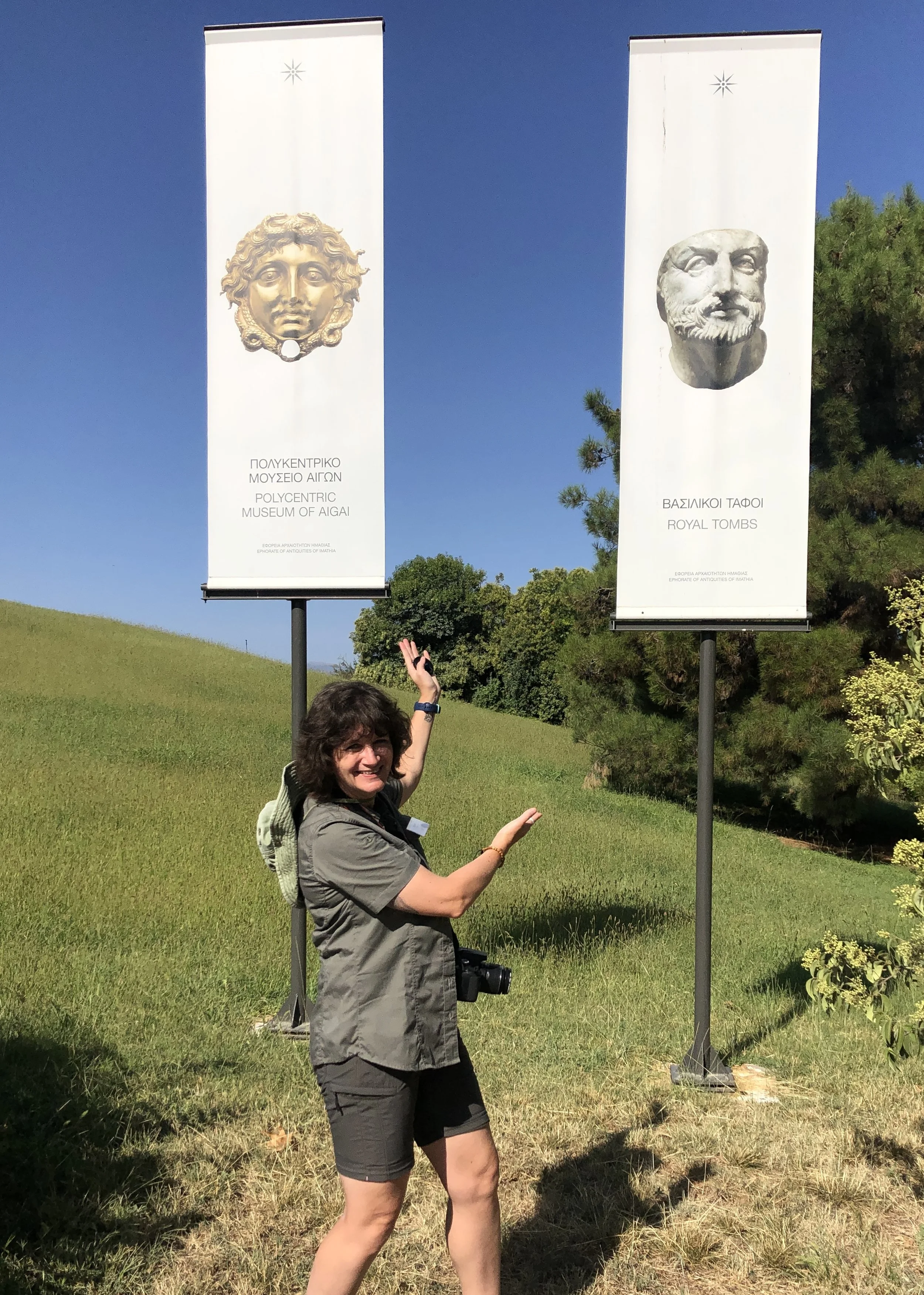 A woman standing outdoors on a grassy hill, smiling and gesturing with her hands, with two tall banners behind her featuring ancient Greek sculptures and Greek text, under a clear blue sky.