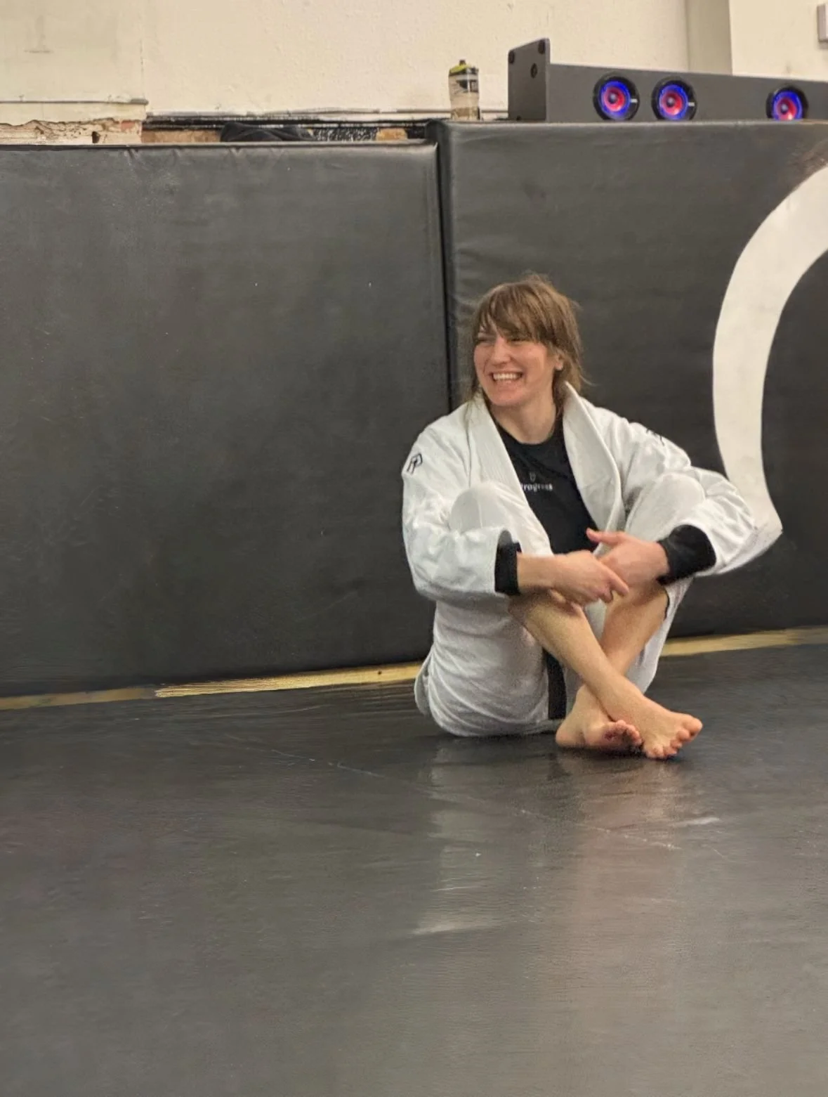 A woman sitting on the floor in a Brazilian Jiu-Jitsu gi, smiling, with a black belt, in front of a black padded wall in a gym or dojo.