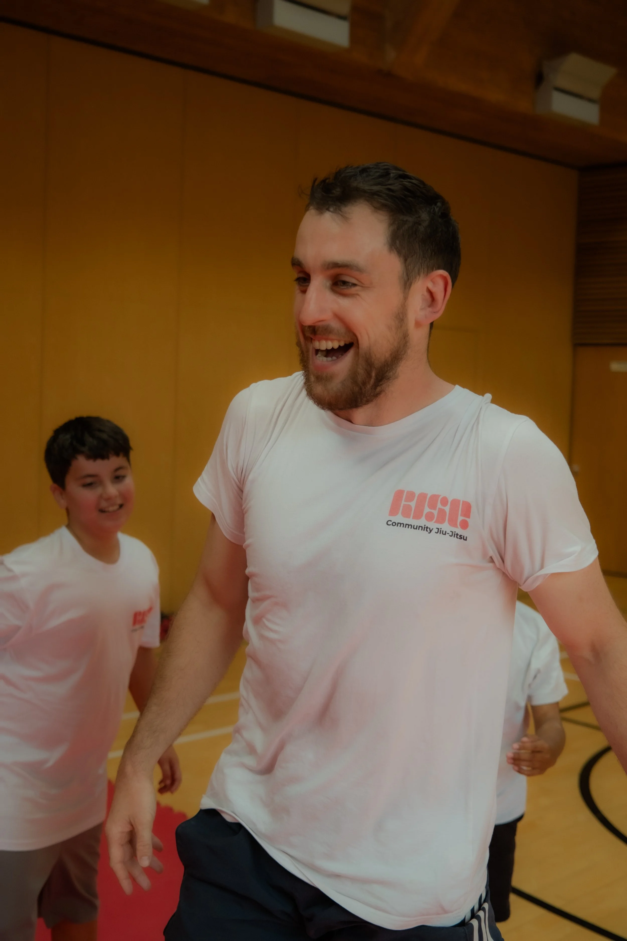 Man smiling and laughing in a gymnasium, wearing a white t-shirt with 'Rise Community Jiu-Jitsu' logo, with children in similar shirts around him.